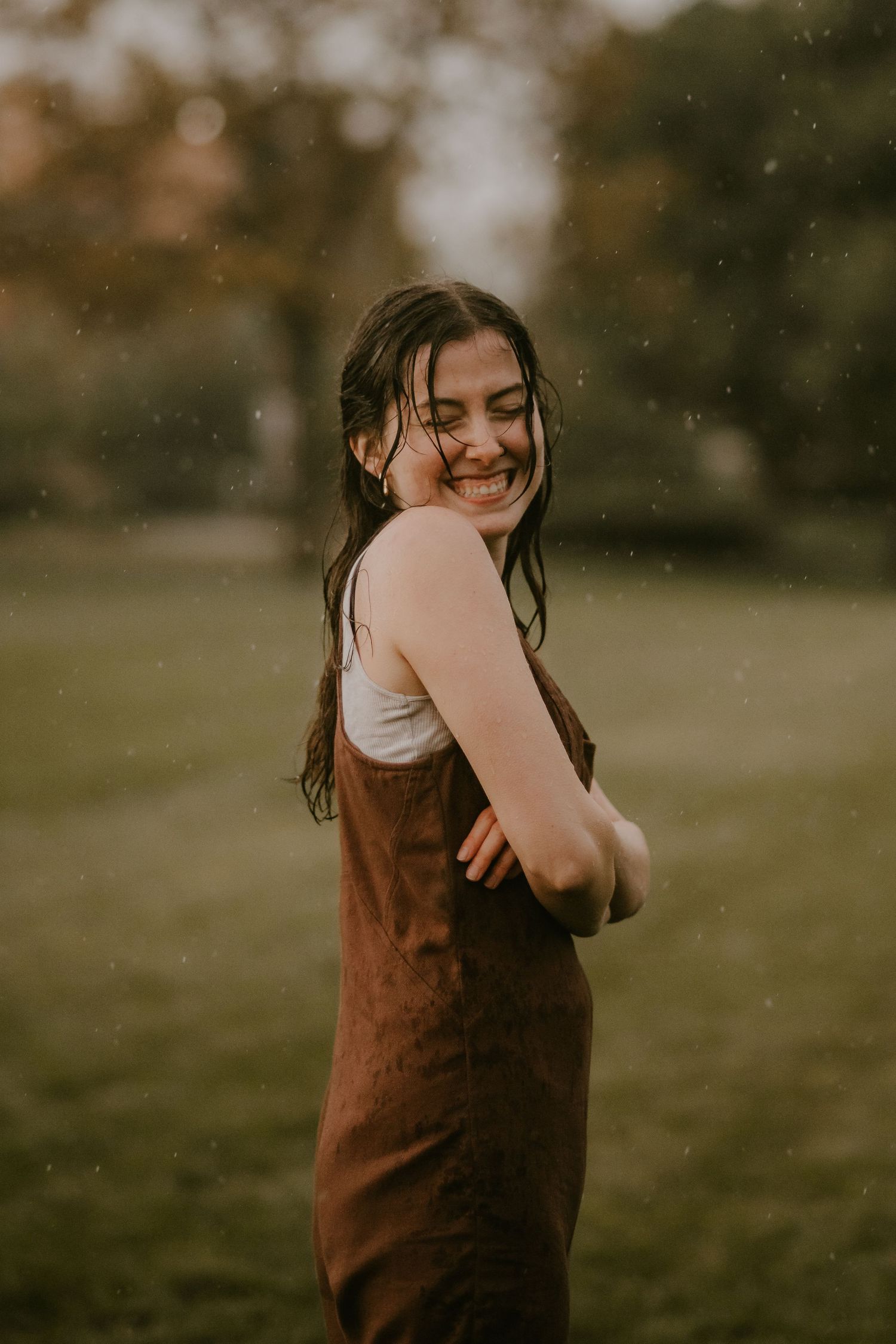 A smiling young woman in a brown apron dress stands in a park, laughing in the rain with wet hair.