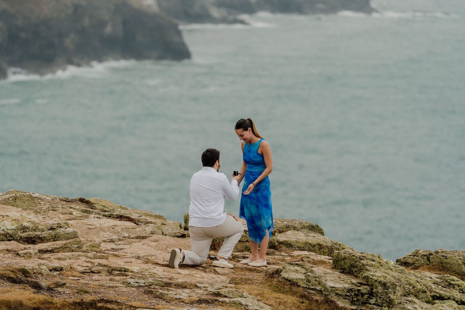 Man proposes to woman in blue dress on the cliff at st agnes head with turquoise ocean backdrop.