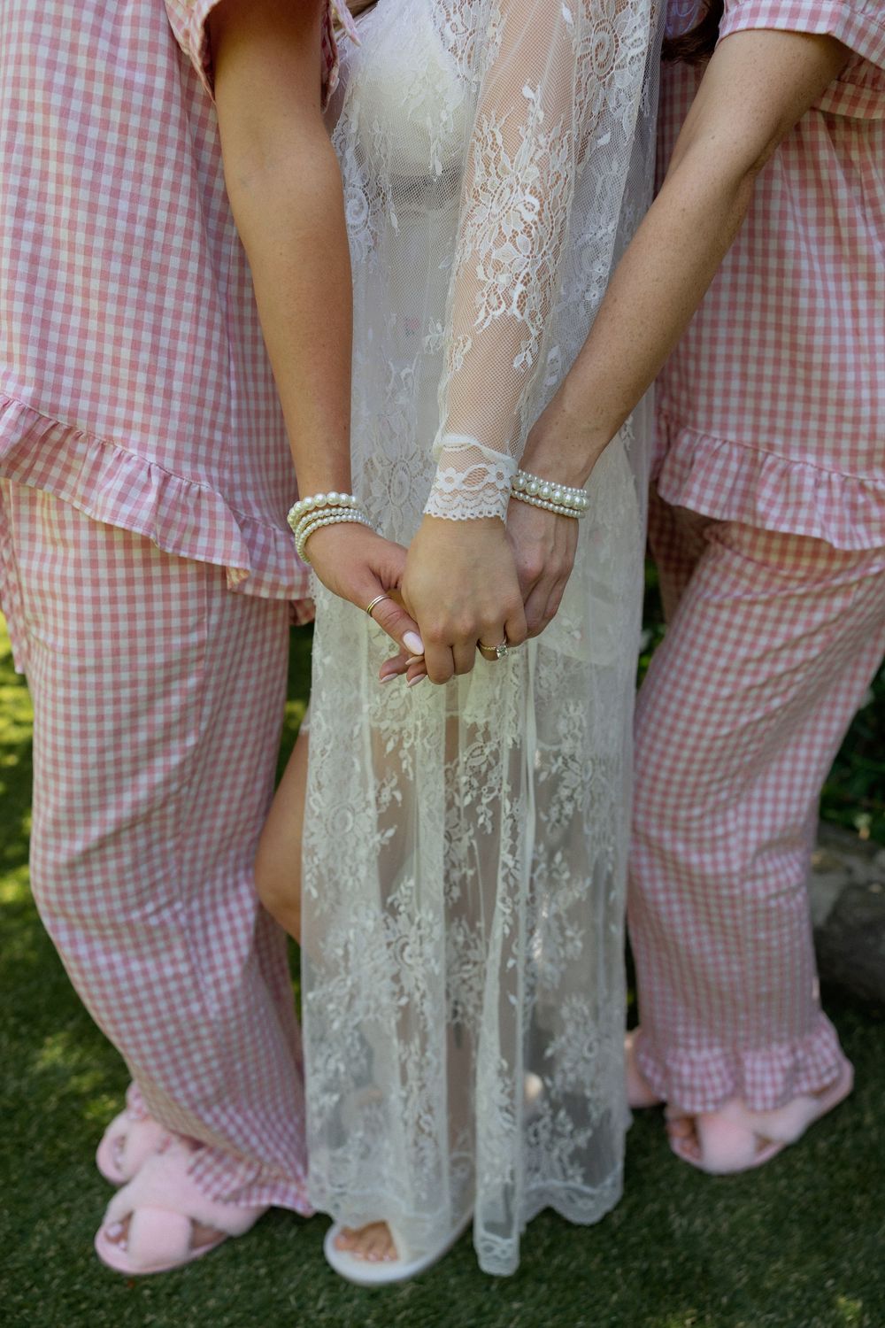 Bride in white lace robe holds hands with bridesmaids wearing pink gingham pajamas outdoors.