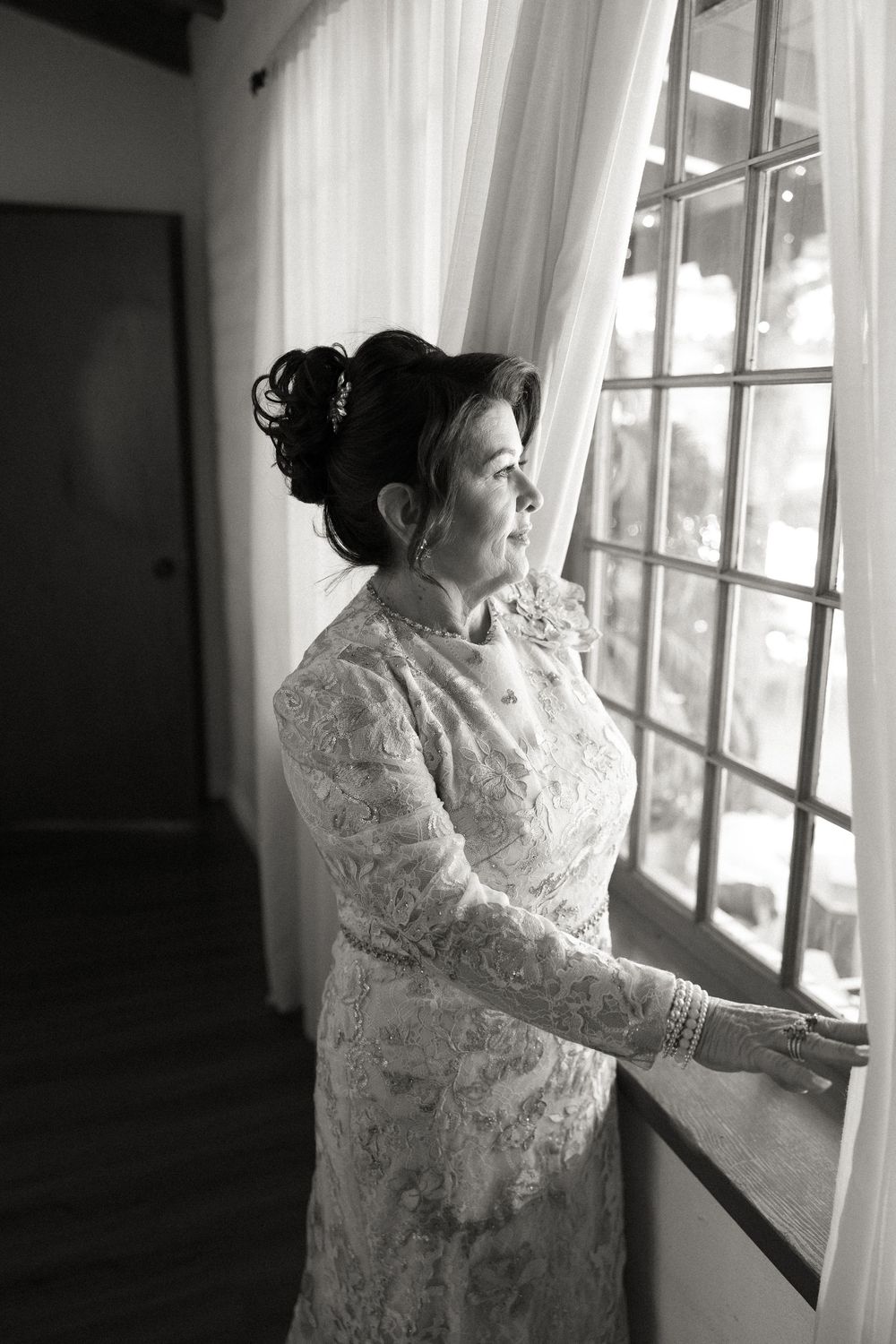 Black and white photo of a bride in lace gown gazing out a window with sheer curtains on her wedding day.