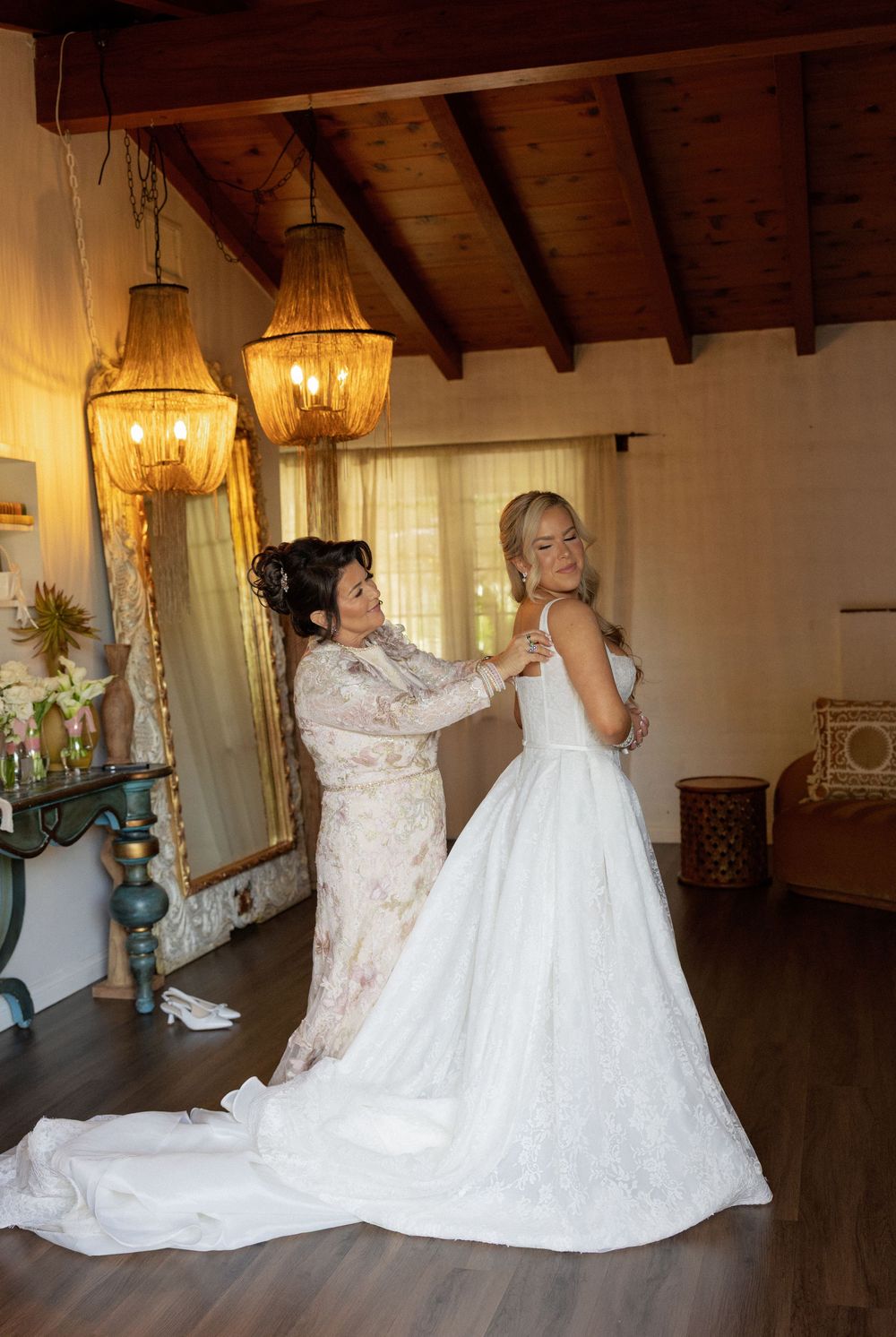 A bride in a white gown gets her dress fastened by a woman in a floral dress inside a rustic room with chandeliers.