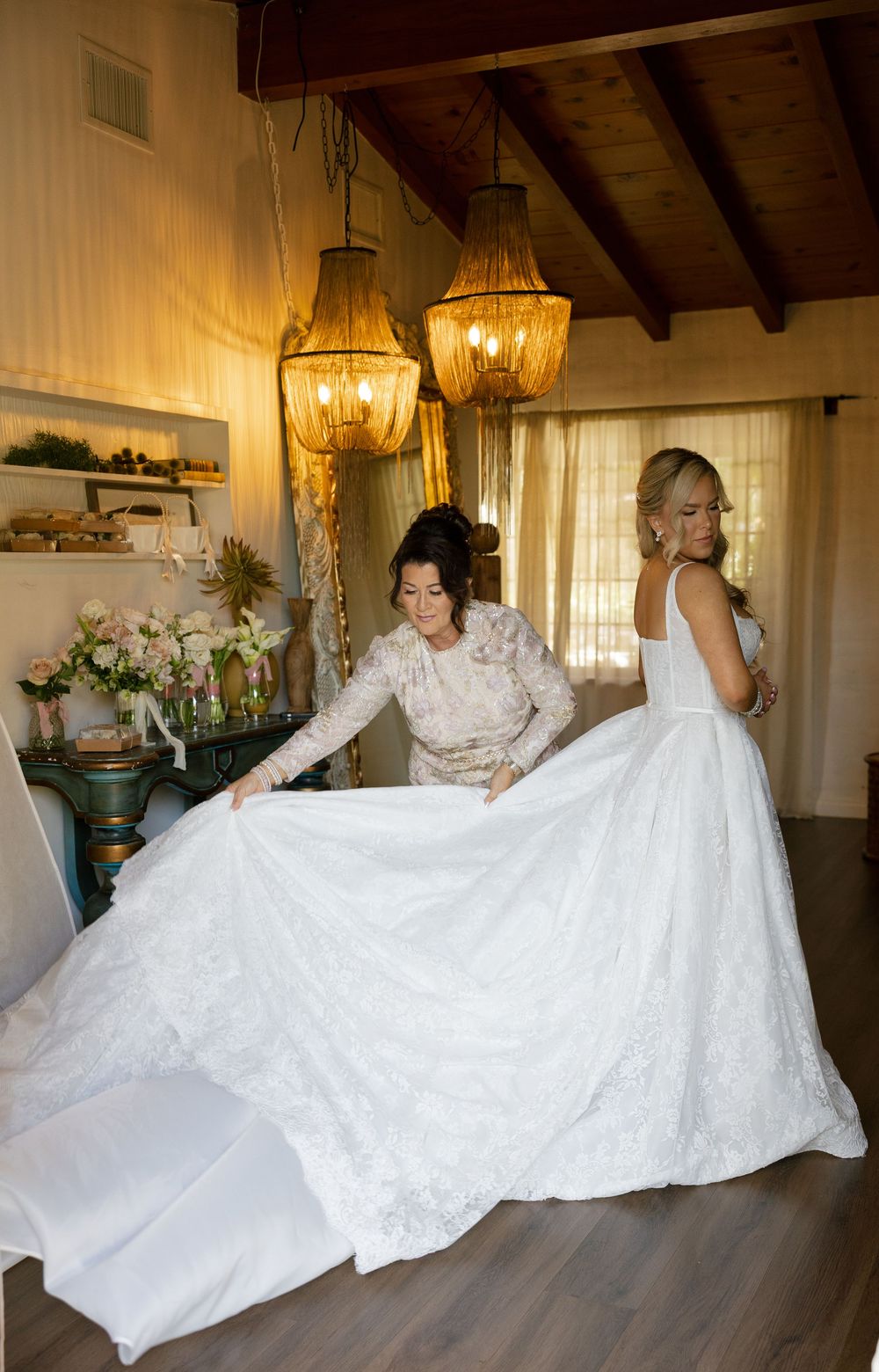 A bride in a white ball gown gets her dress arranged by a helper inside an elegant wood-beamed room with chandelier lighting.