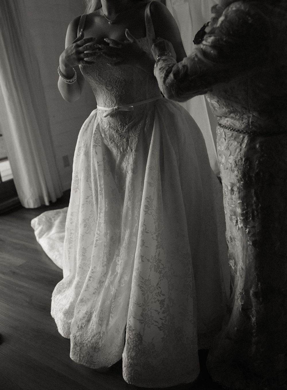 Black and white photo of a bride in an elegant lace wedding gown being helped with her dress before the ceremony.