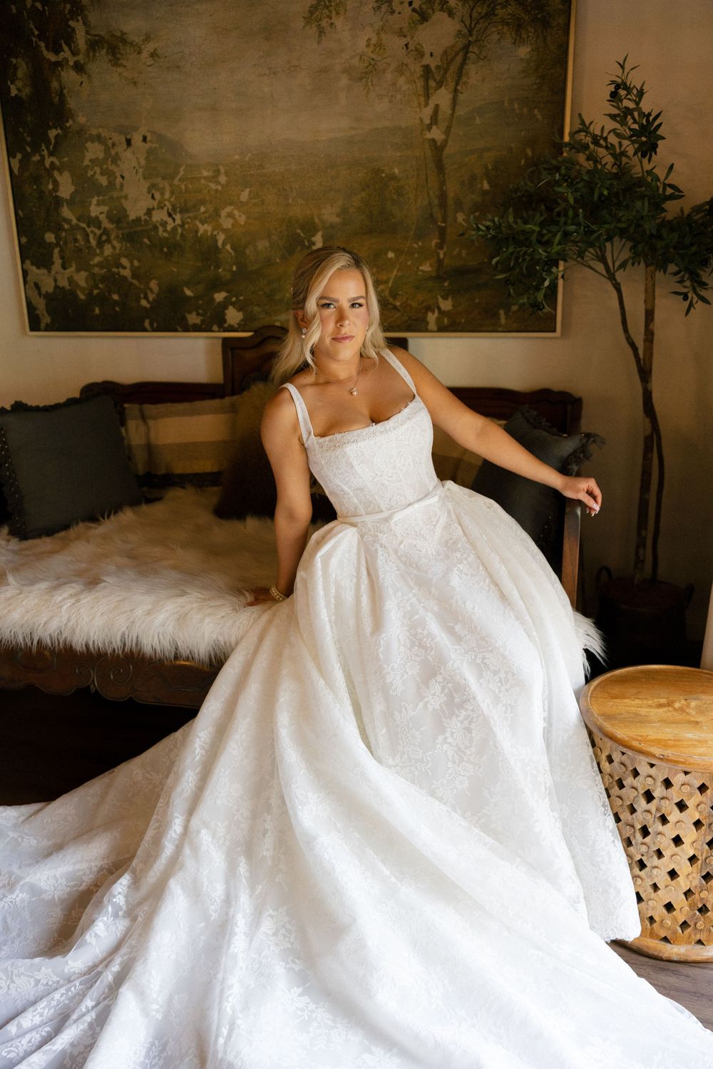 Bride in a stunning white lace ballgown wedding dress seated elegantly in a warmly decorated room.