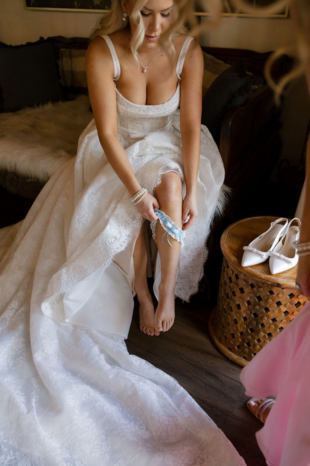 Bride in a white lace wedding gown putting on a blue garter while seated, with heels nearby.
