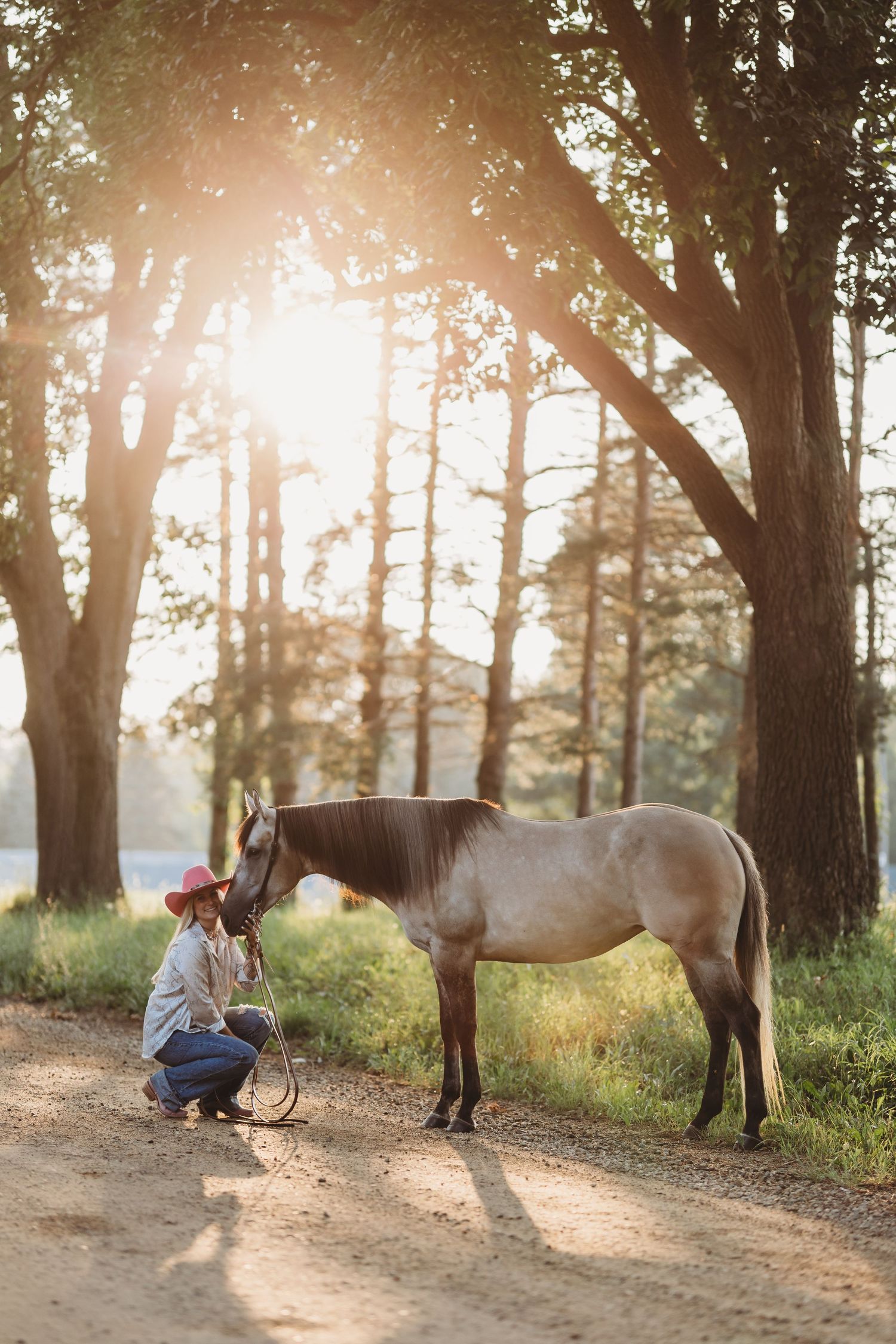 Woman in pink hat crouches to bond with a buckskin horse on a sunlit forest path at golden hour.