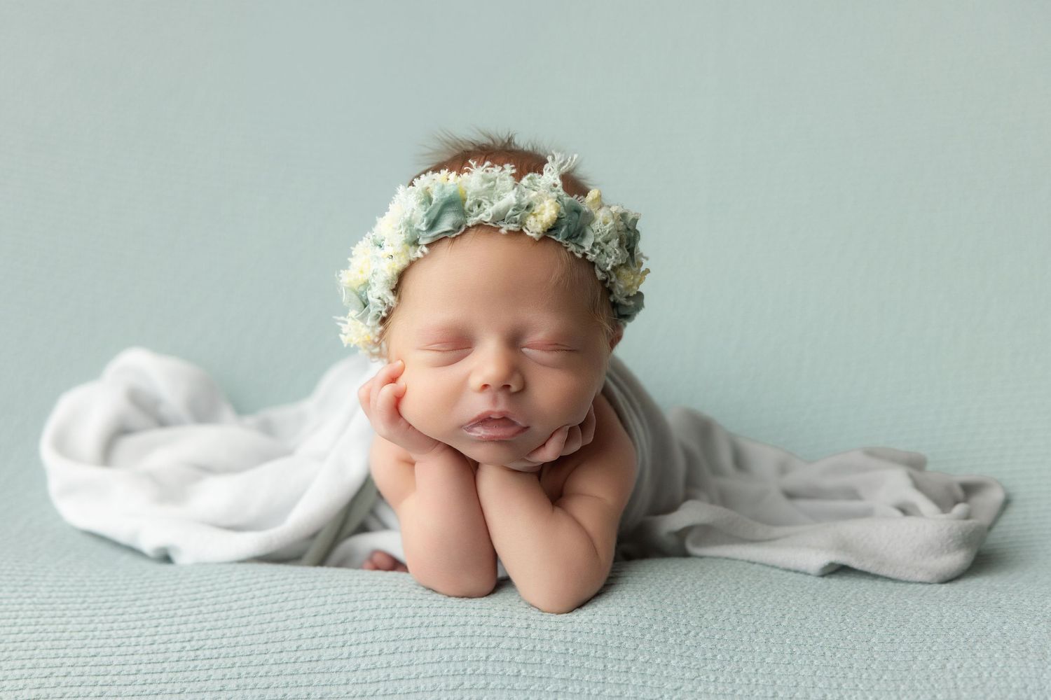 Newborn baby resting chin on hands inside a teal bucket with soft blanket, studio photography.
