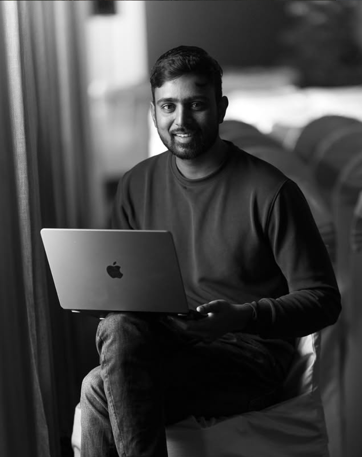 Young South Asian man smiling while holding an Apple MacBook laptop, black and white portrait photo.
