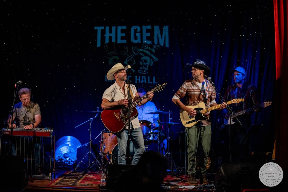 Band performing live at The Gem Hall with guitarist, keyboardist, and drummer on a decorated stage under blue lights.