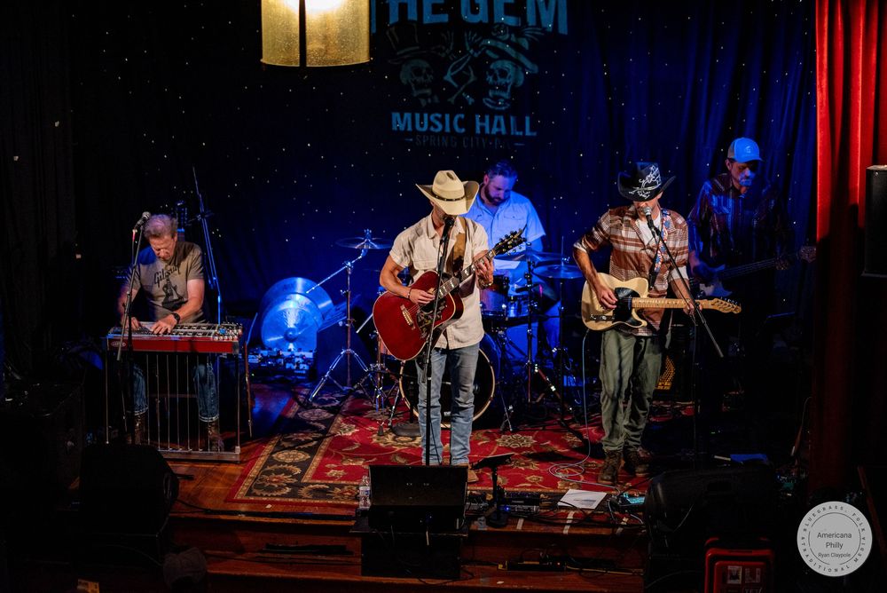 Live band performing on stage at The Gem Music Hall, featuring guitarist in cowboy hat, keyboardist, and guitarist under warm lights.