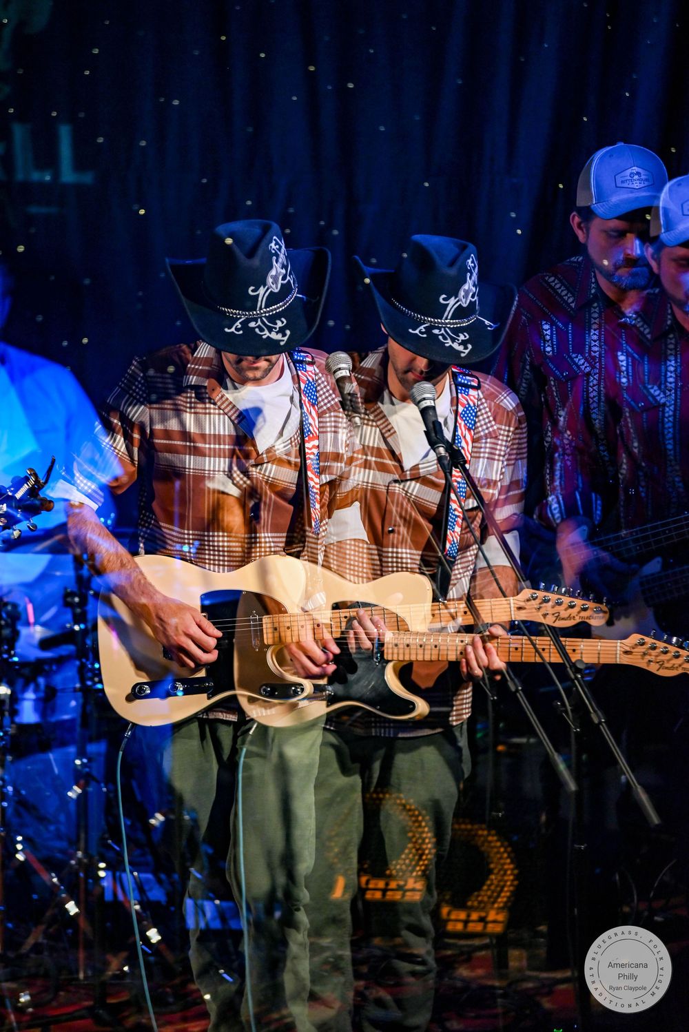 Two cowboys in matching hats and plaid shirts play guitars together on a country music stage.