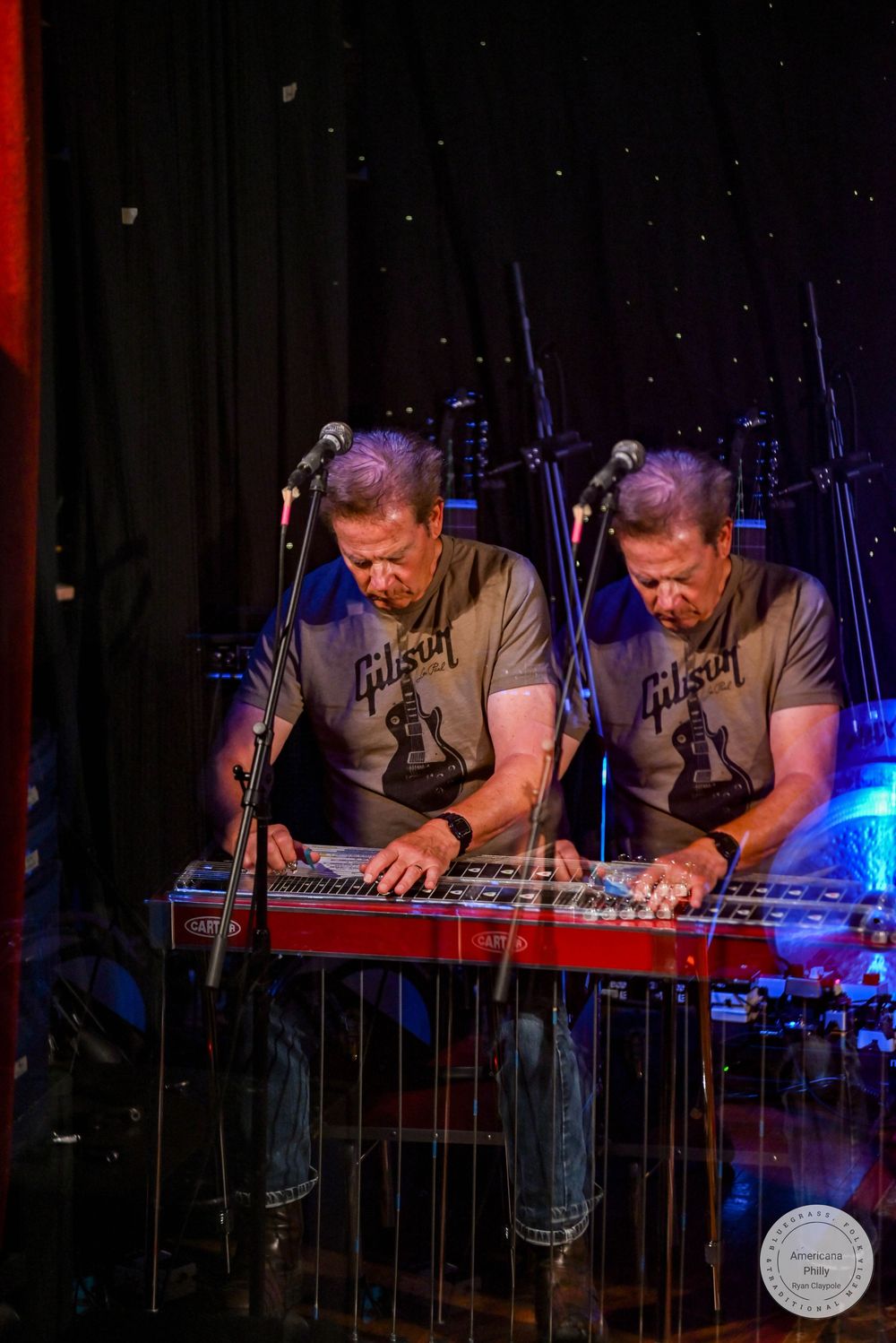 Two musicians in Gibson t-shirts play pedal steel guitars on a dimly lit stage with microphones.