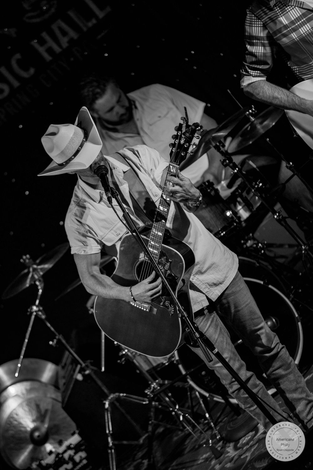 Black and white photo of a guitarist wearing a cowboy hat performing on stage at a music hall with a drummer behind him.