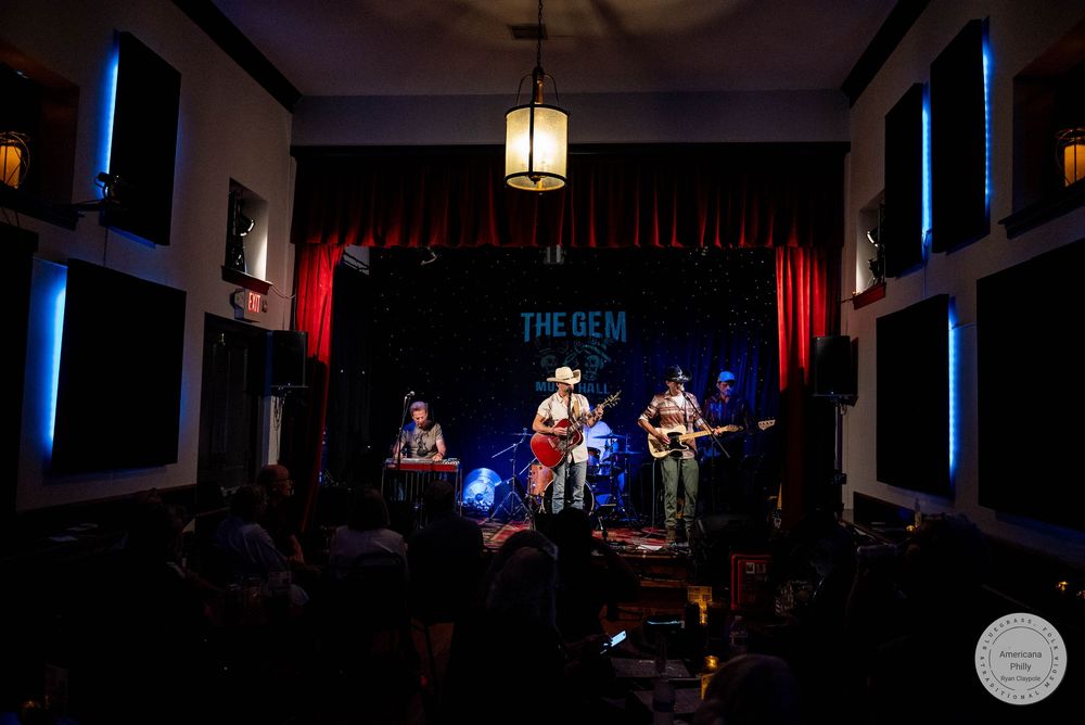 Live band performing on stage at The Gem venue, with red curtains and blue lighting.