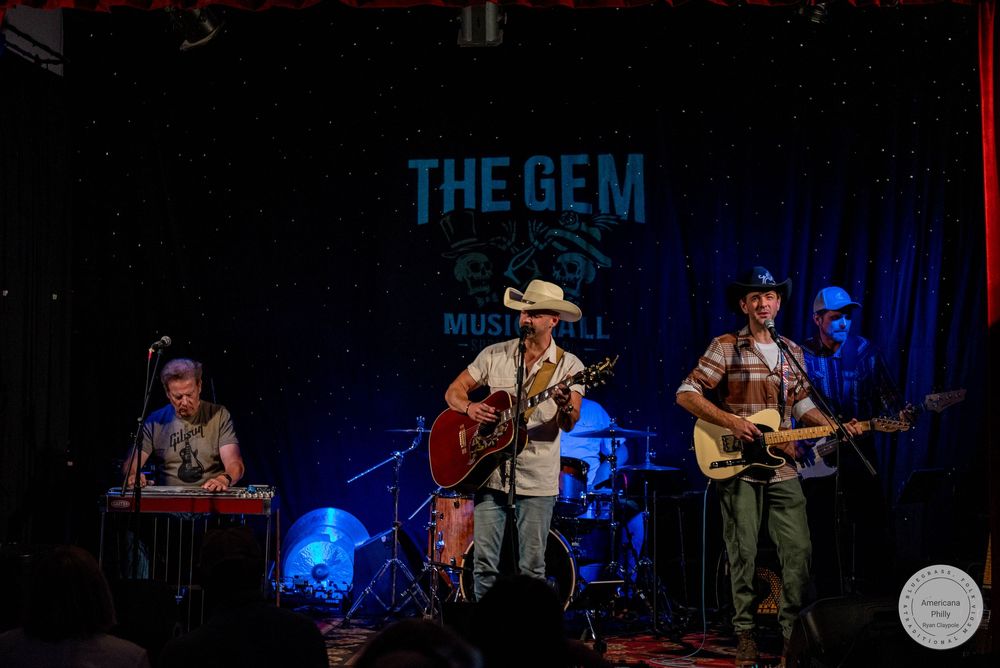 Three musicians perform live on stage at The Gem Music venue, featuring guitars and keyboards under warm stage lighting.