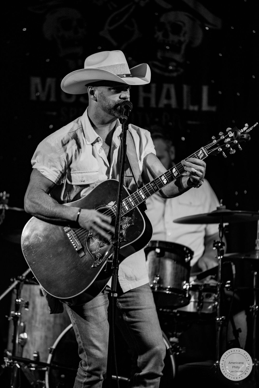 Country musician in white cowboy hat playing acoustic guitar on stage at a music hall in black and white photo.