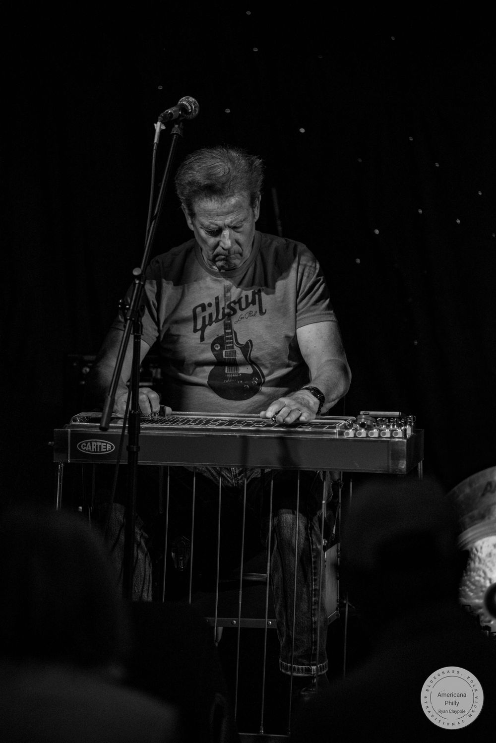 Man in Gibson t-shirt playing pedal steel guitar on stage at a live music venue, black and white photo.