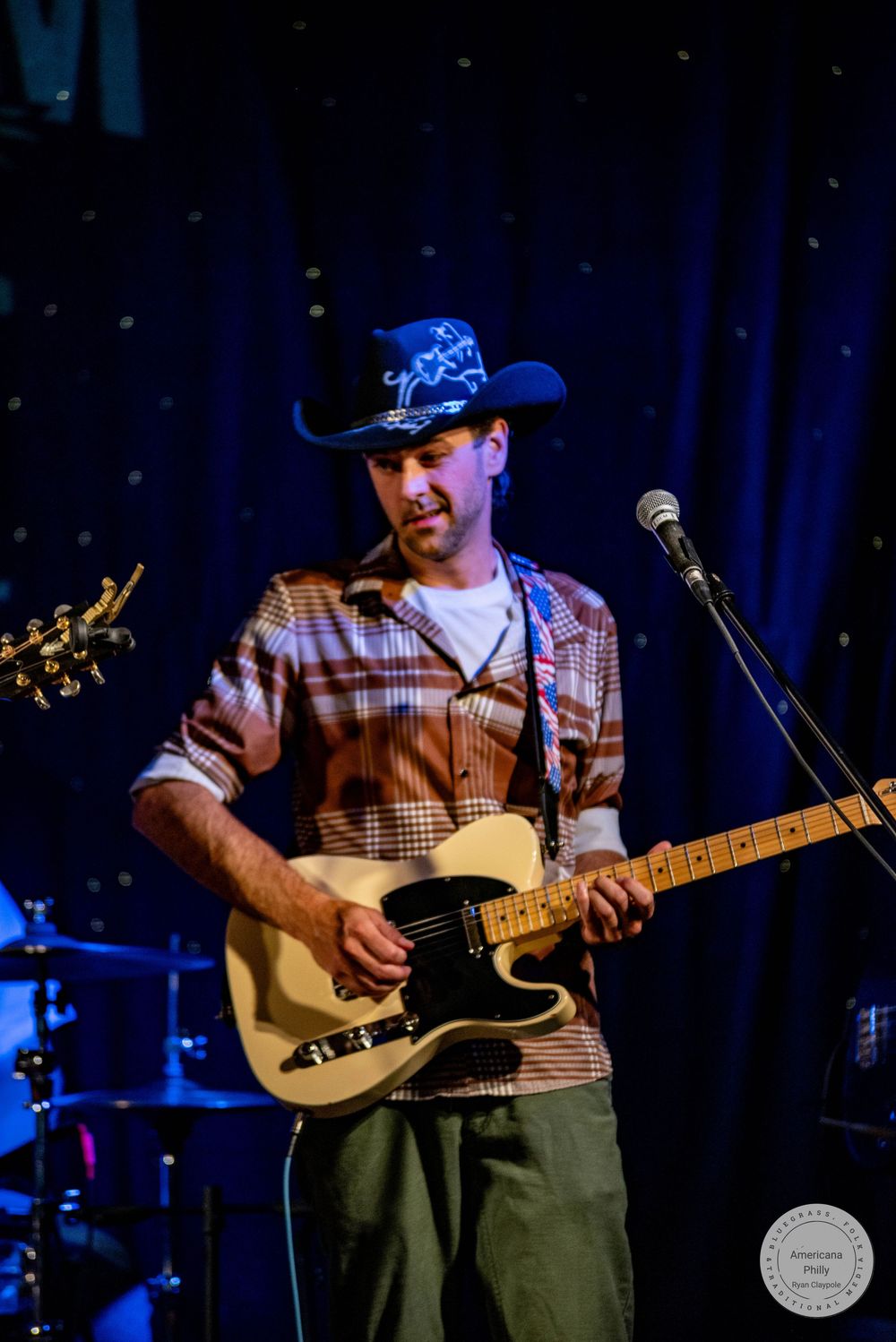 A male guitarist wearing a cowboy hat performs on stage with a Telecaster guitar near a microphone under blue stage lighting.