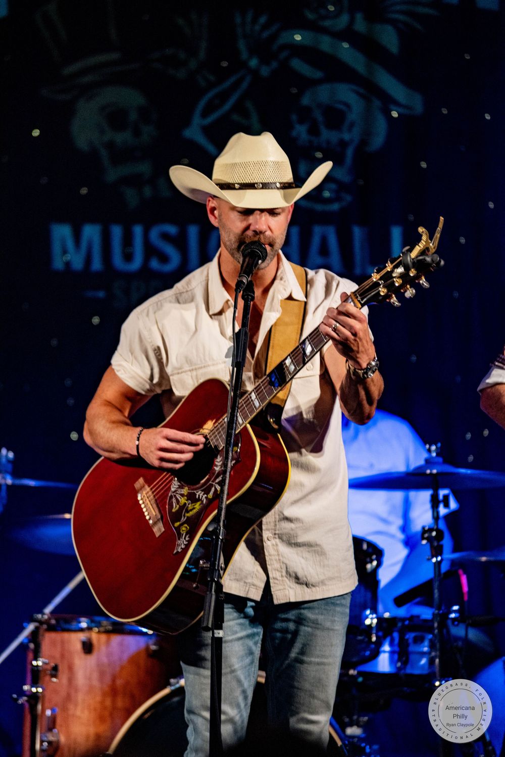 Country musician in white cowboy hat plays red acoustic guitar on stage at Music Hall venue.