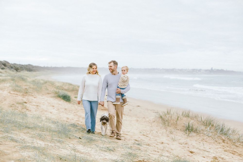 Family with dog walking on beach dunes, parents holding toddler, coastal scenery in background.