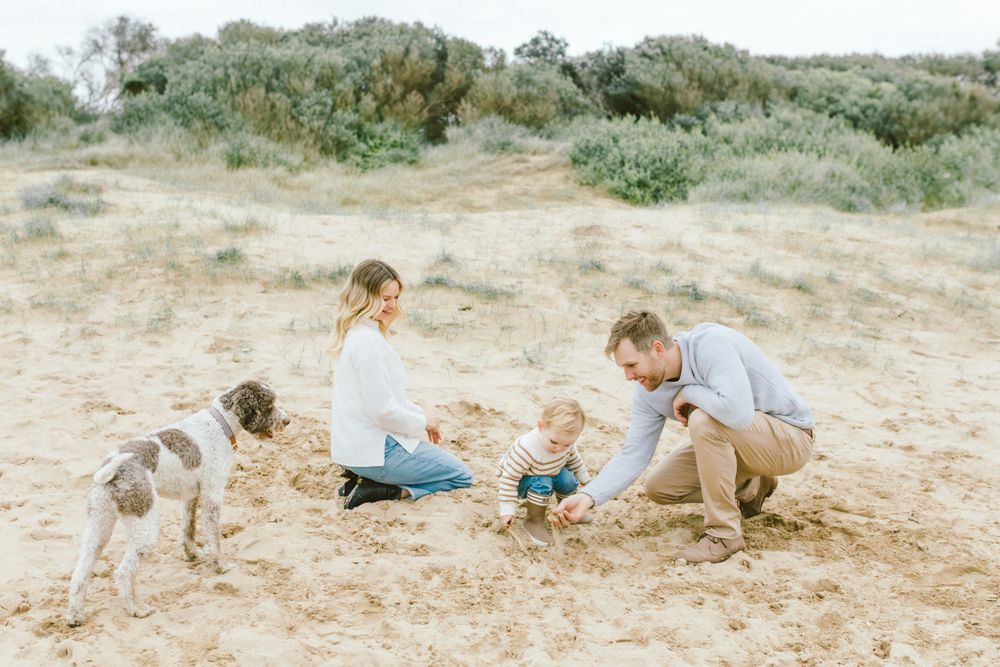 Family with dog playing on sandy beach, parents and toddler enjoying outdoor time together in natural coastal setting.