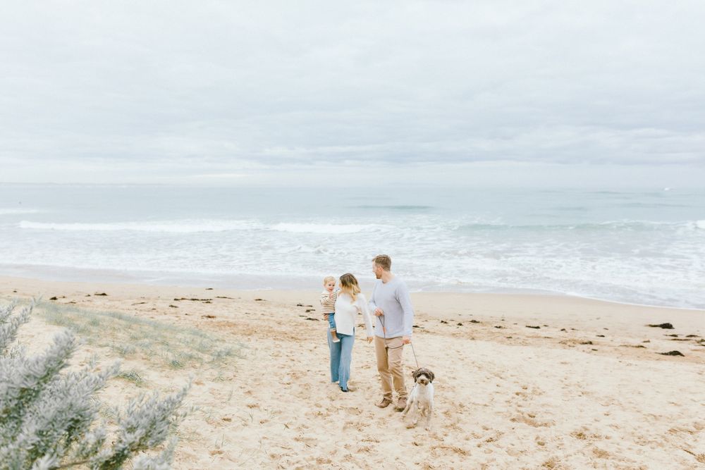 Couple with dog standing on sandy beach near ocean waves under cloudy sky.