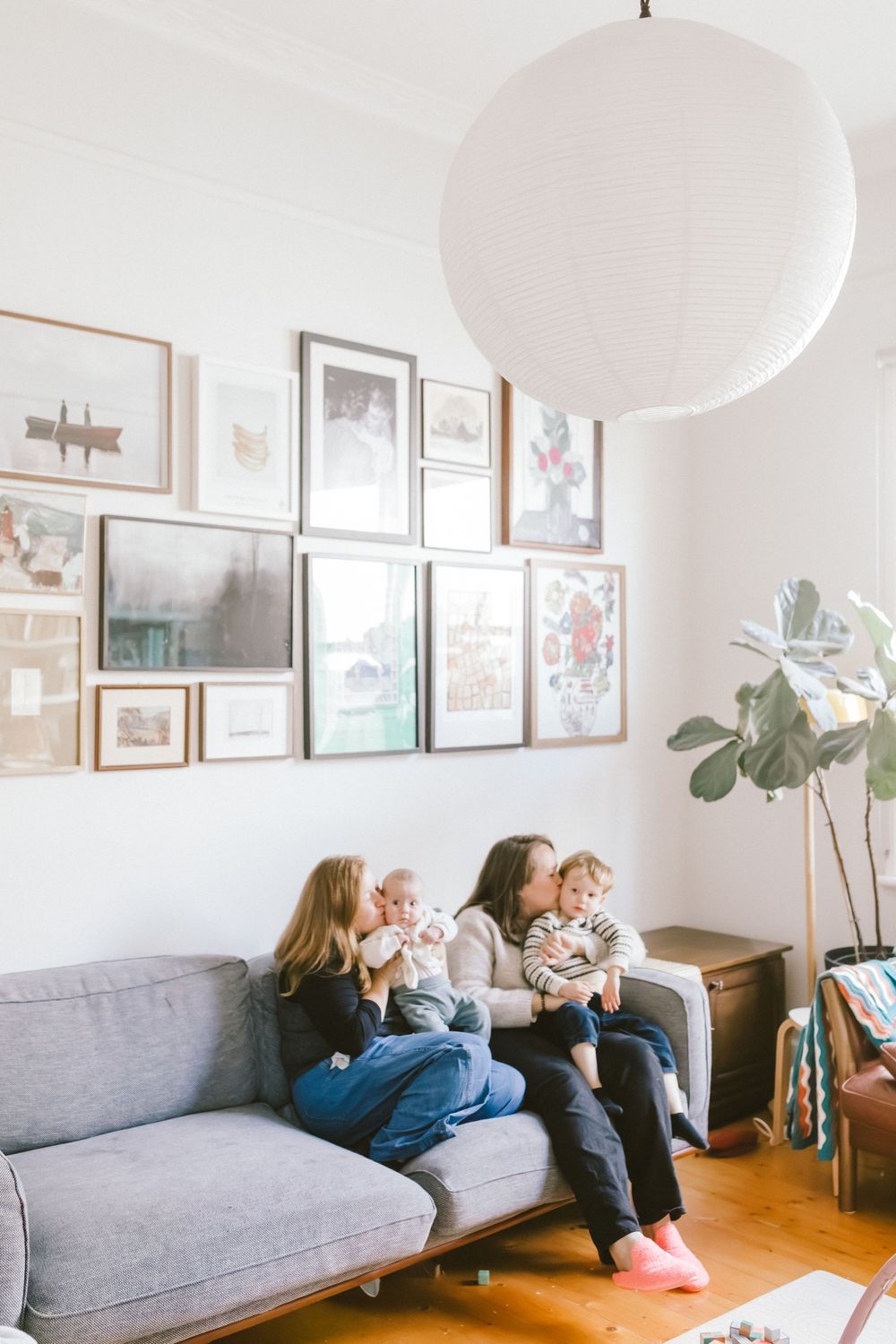 Two women sit on a gray sofa holding babies, surrounded by gallery wall art and a white pendant light.