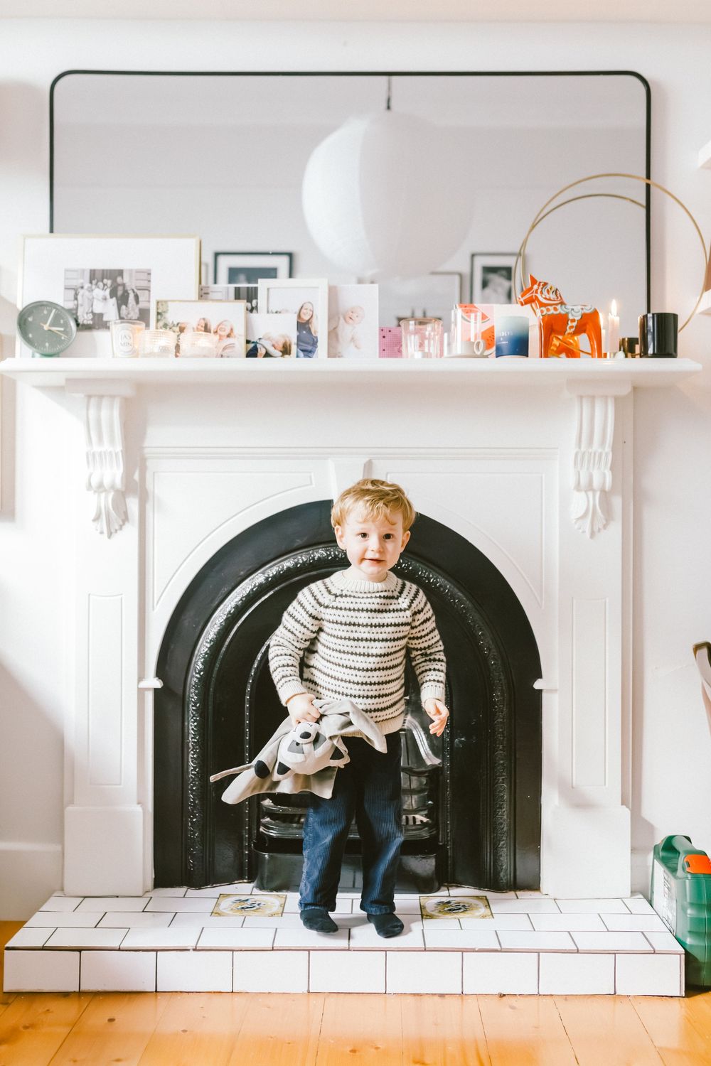 Toddler boy in striped sweater standing in a white fireplace holding a stuffed animal toy in a cozy living room.