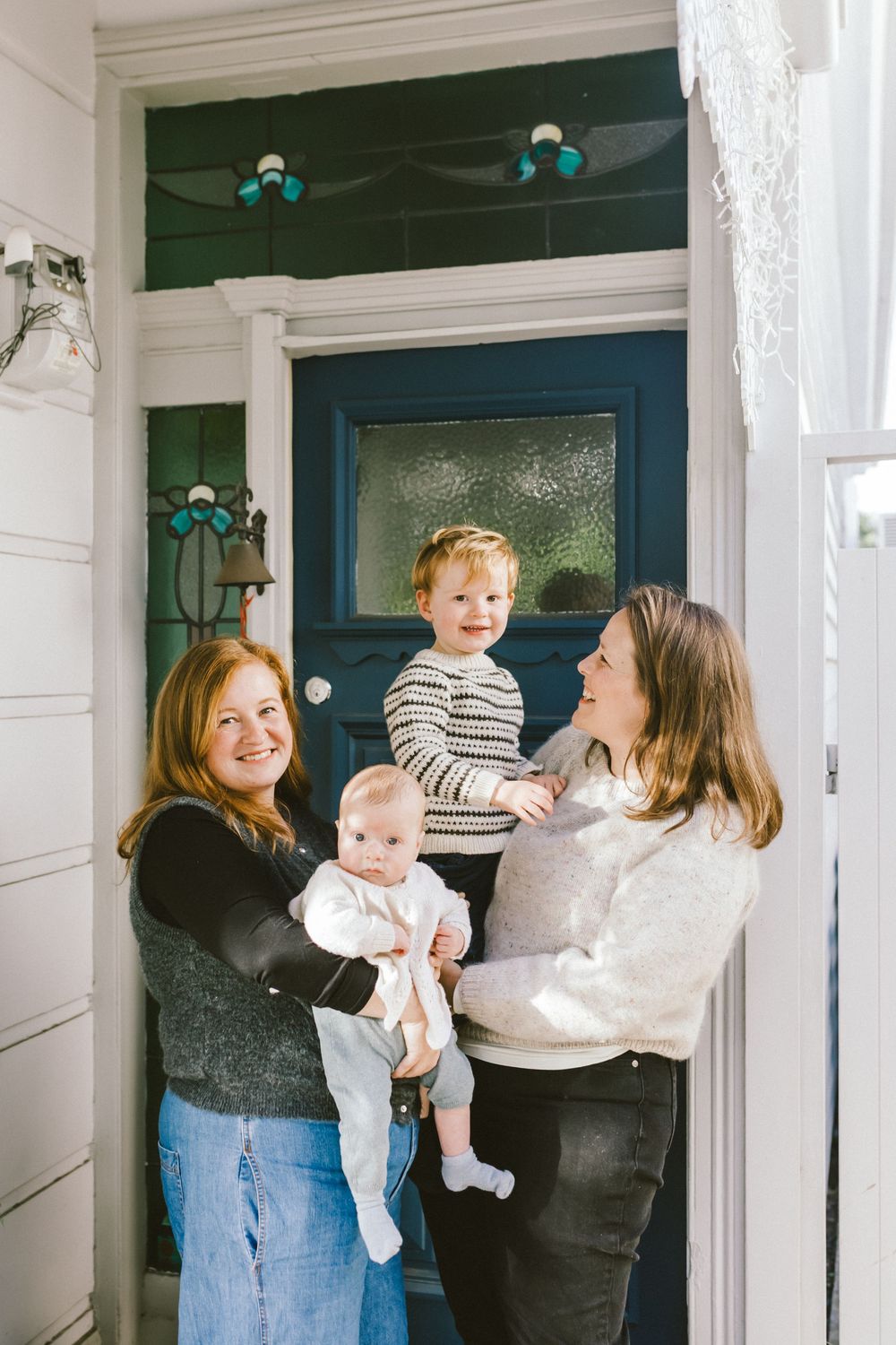 Two women smiling at front door, one holding a baby and the other holding a toddler in a striped shirt.
