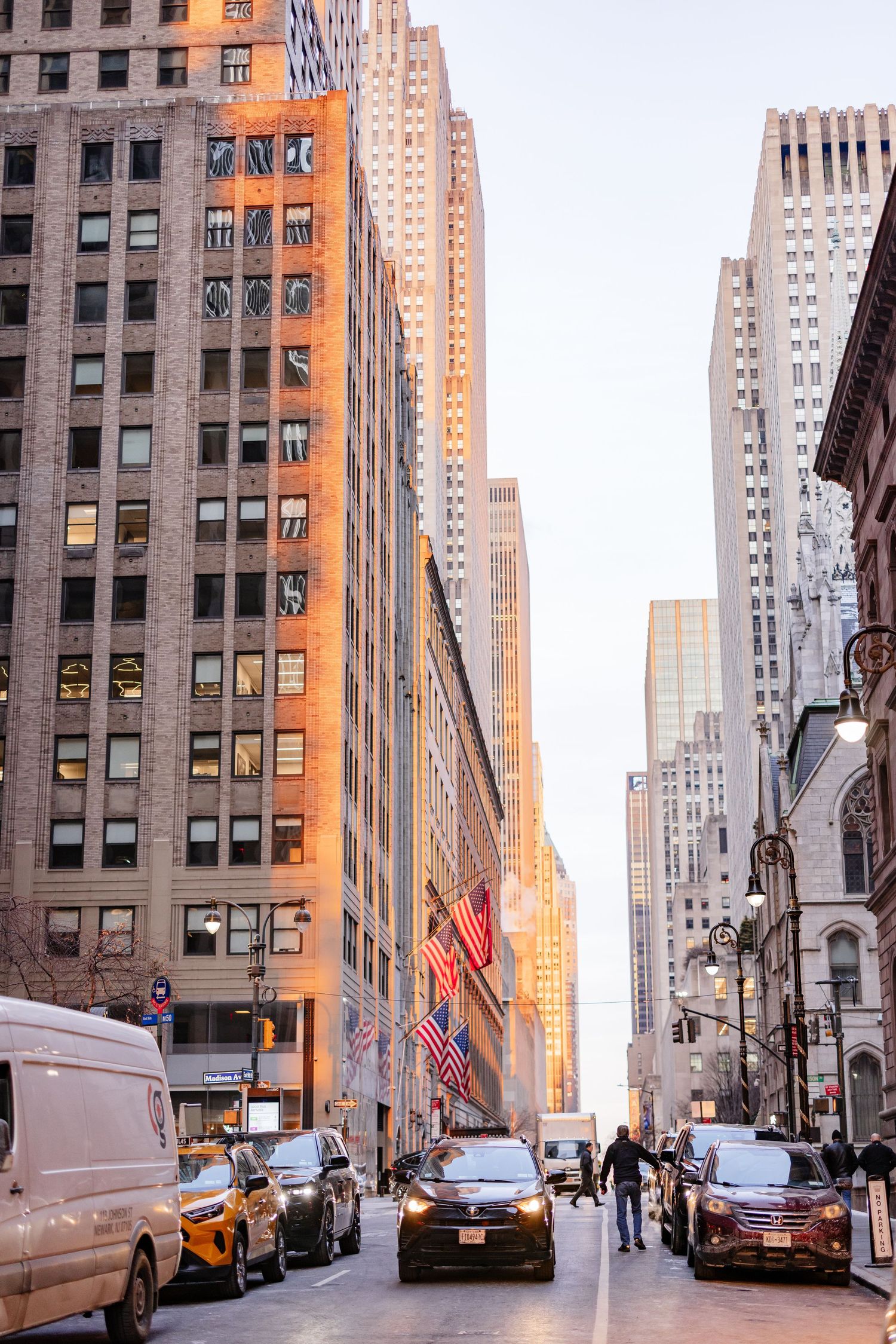 Golden hour sunlight illuminates NYC skyscrapers on a busy Manhattan street with American flags and city traffic.