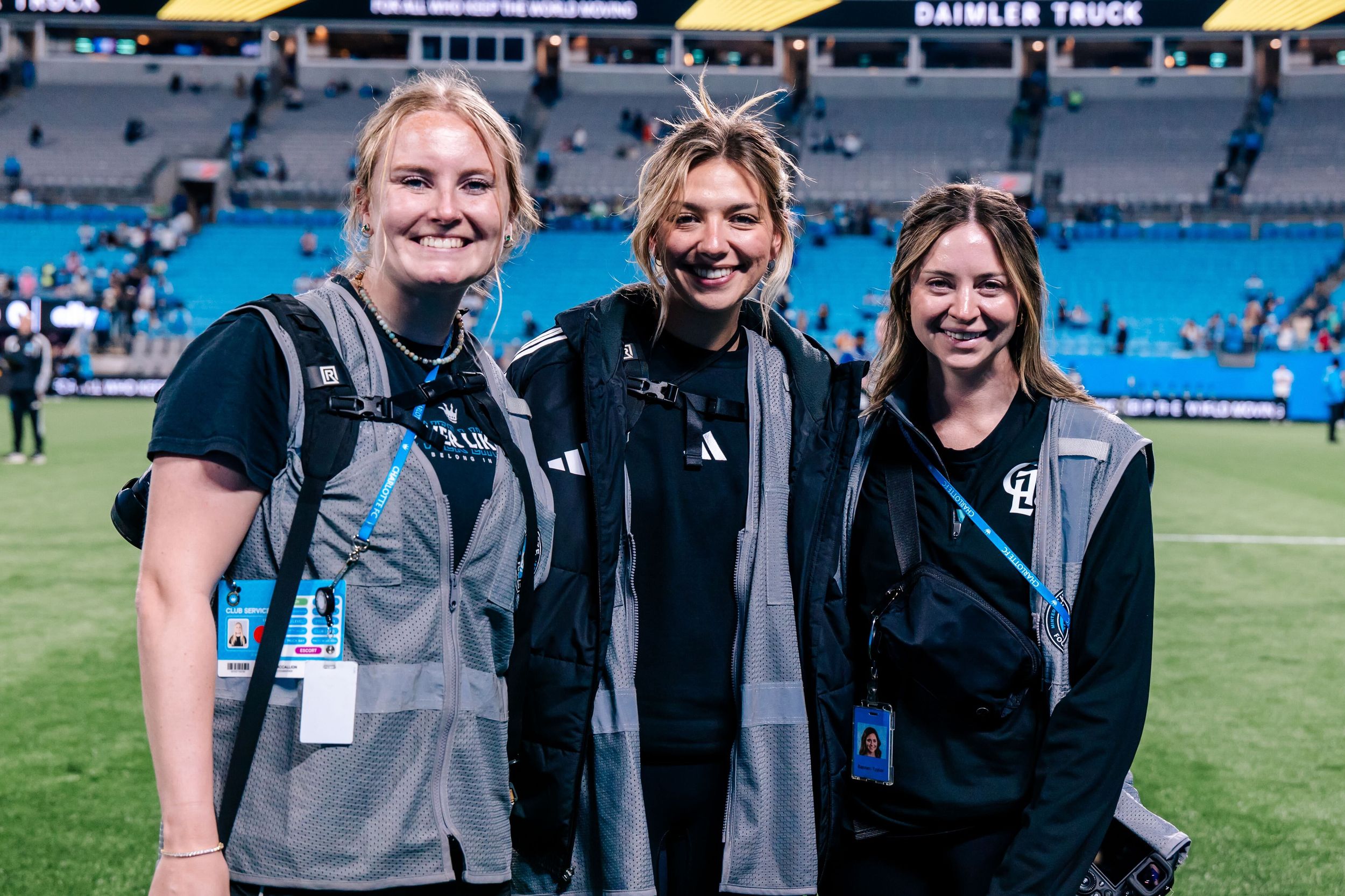 Three smiling women in black uniforms with lanyards pose on a football field with stadium lights in the background.