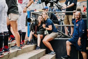 Two female photographers smile while crouching on stadium steps during a sporting event.