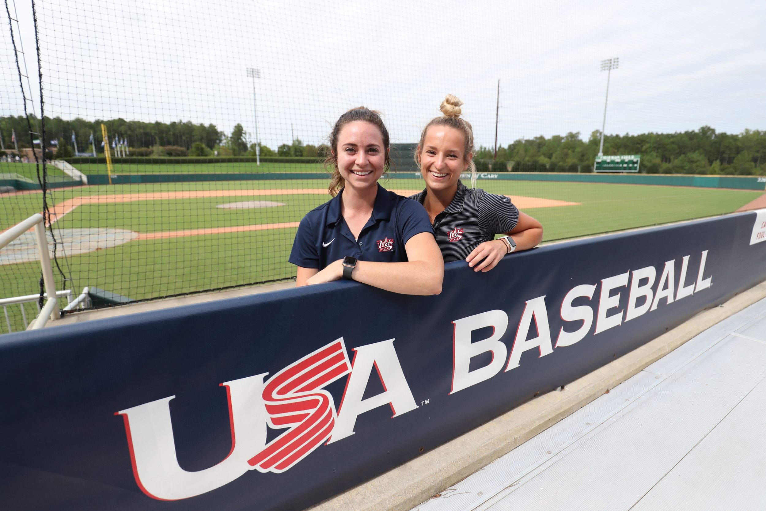 Two women posing behind a USA Baseball dugout sign at an outdoor baseball stadium.