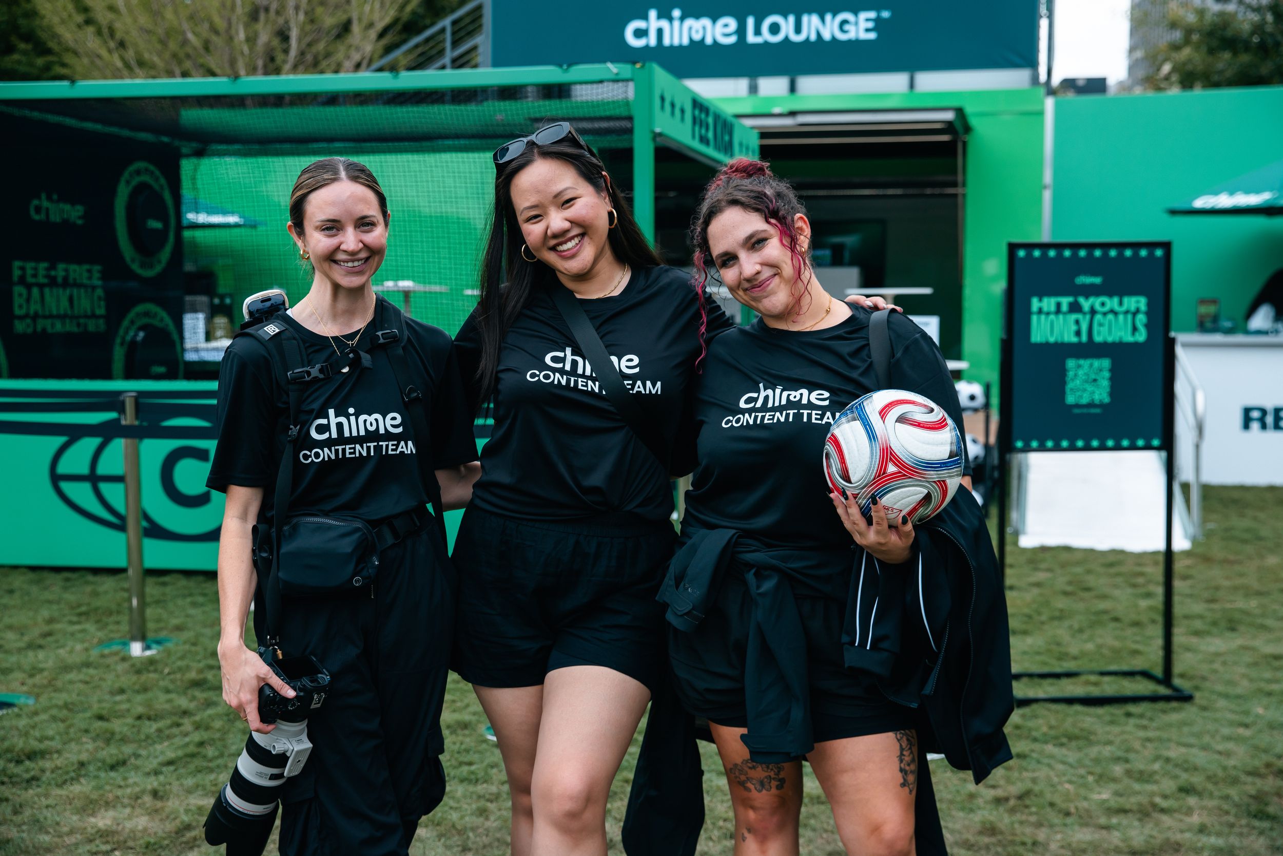 Three women in Chime Content Team shirts smile at a Chime Lounge event, one holding a soccer ball.