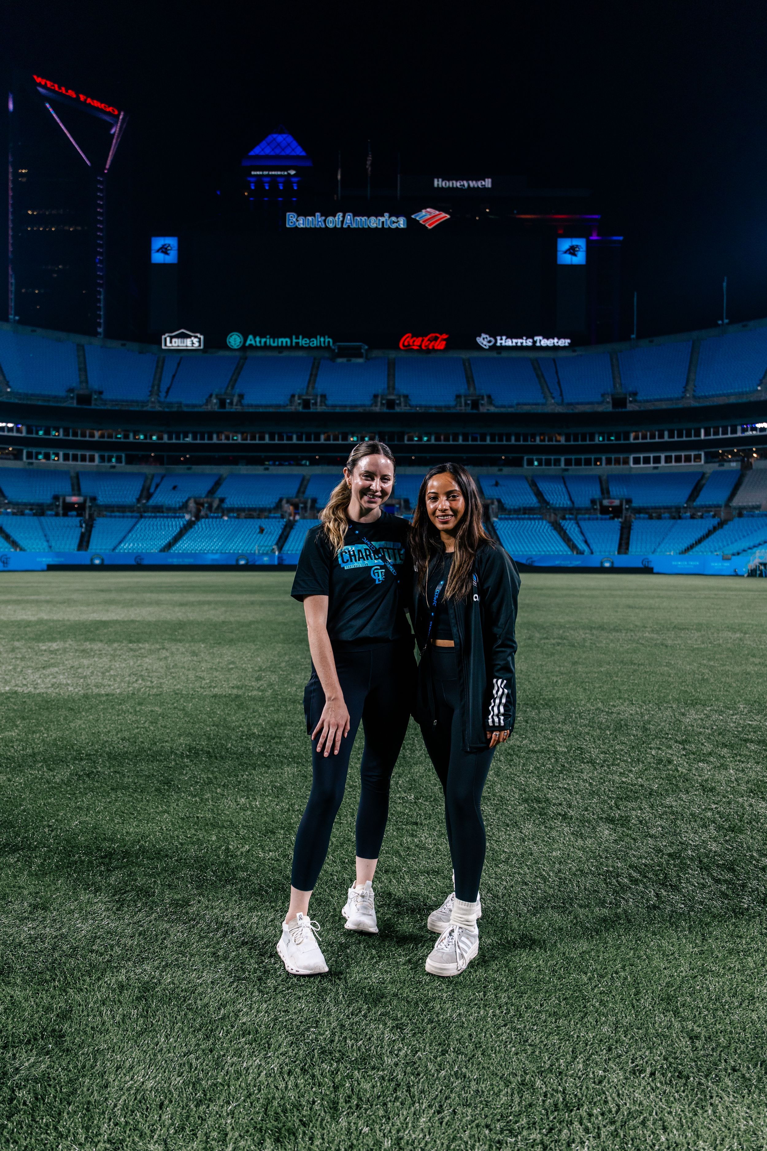 Two women smile together on football field at night inside Bank of America Stadium.