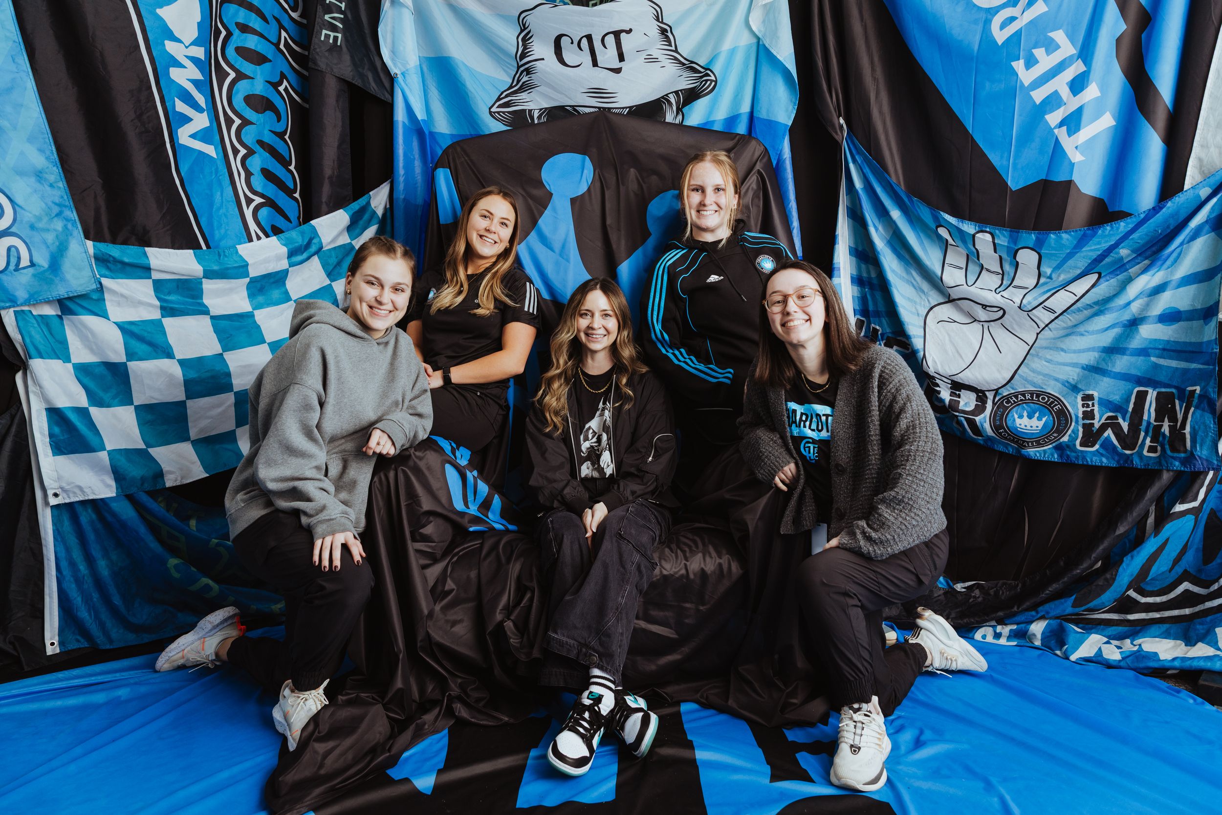 Five women pose together smiling among blue and black soccer fan flags and banners.