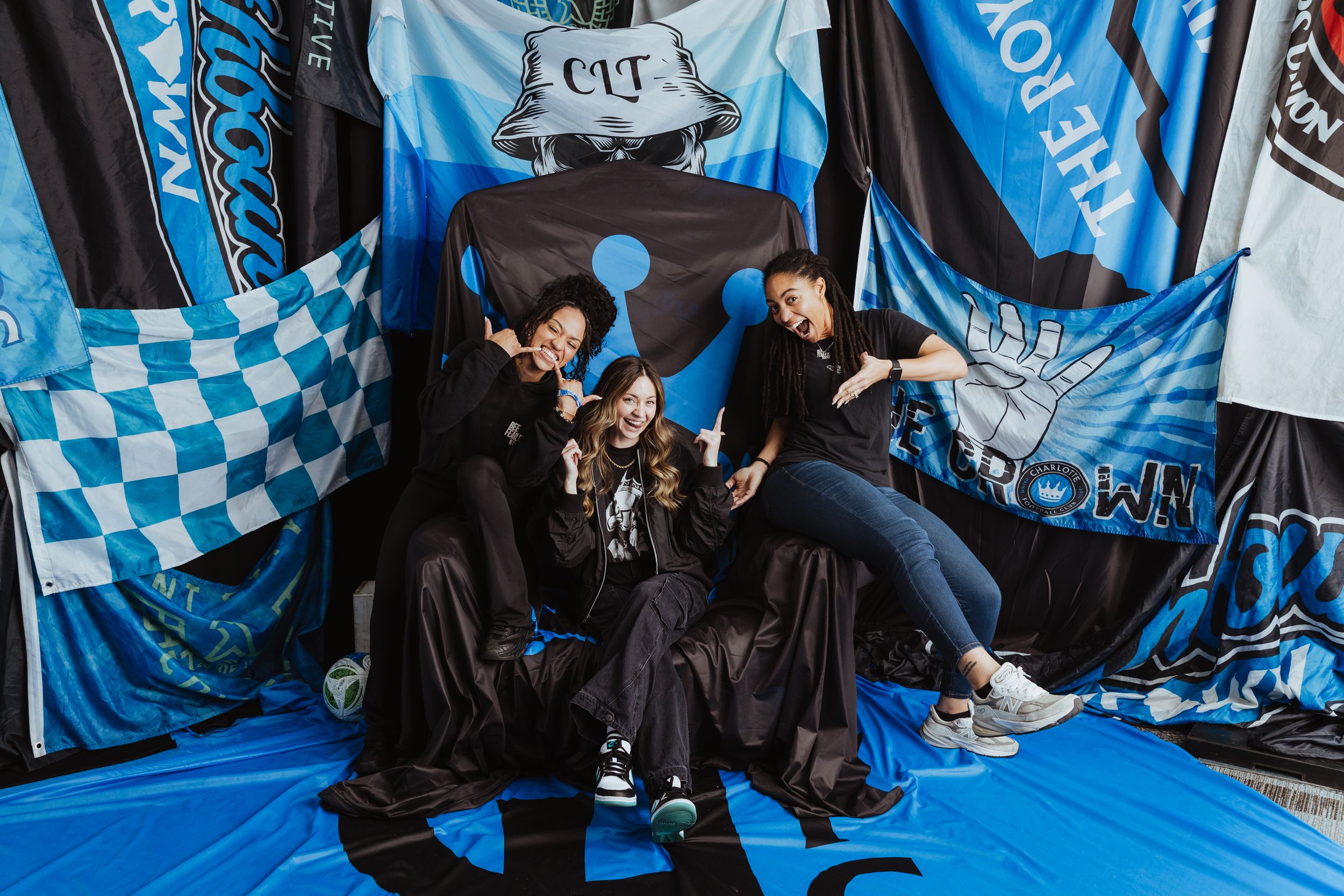 Three women pose playfully among blue and black soccer supporter flags and banners.