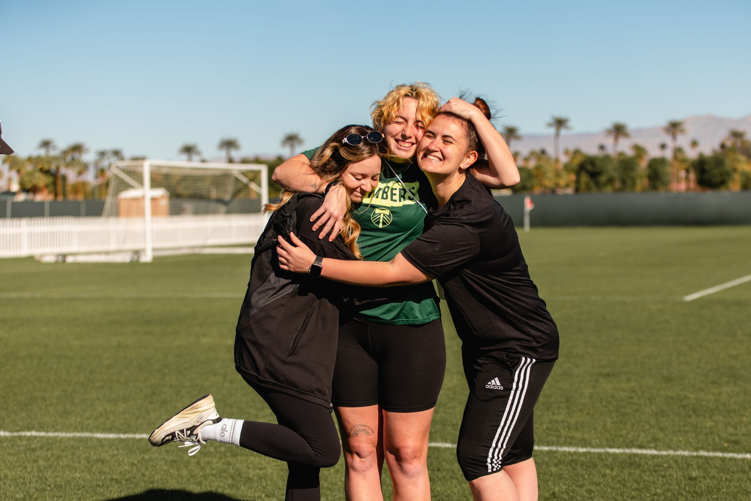 Three women share a group hug on a green soccer field with palm trees behind.