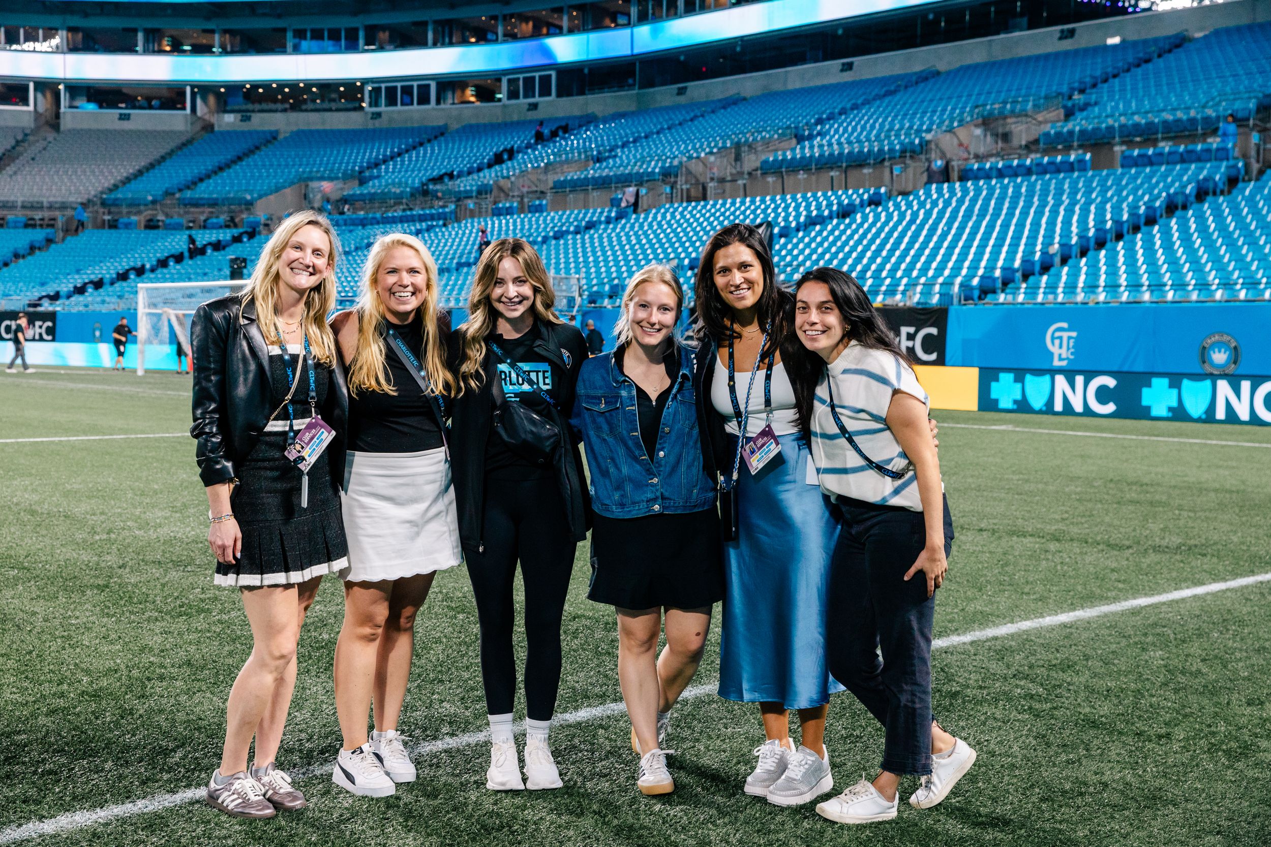 Seven women pose together smiling on the field inside a soccer stadium at an event.