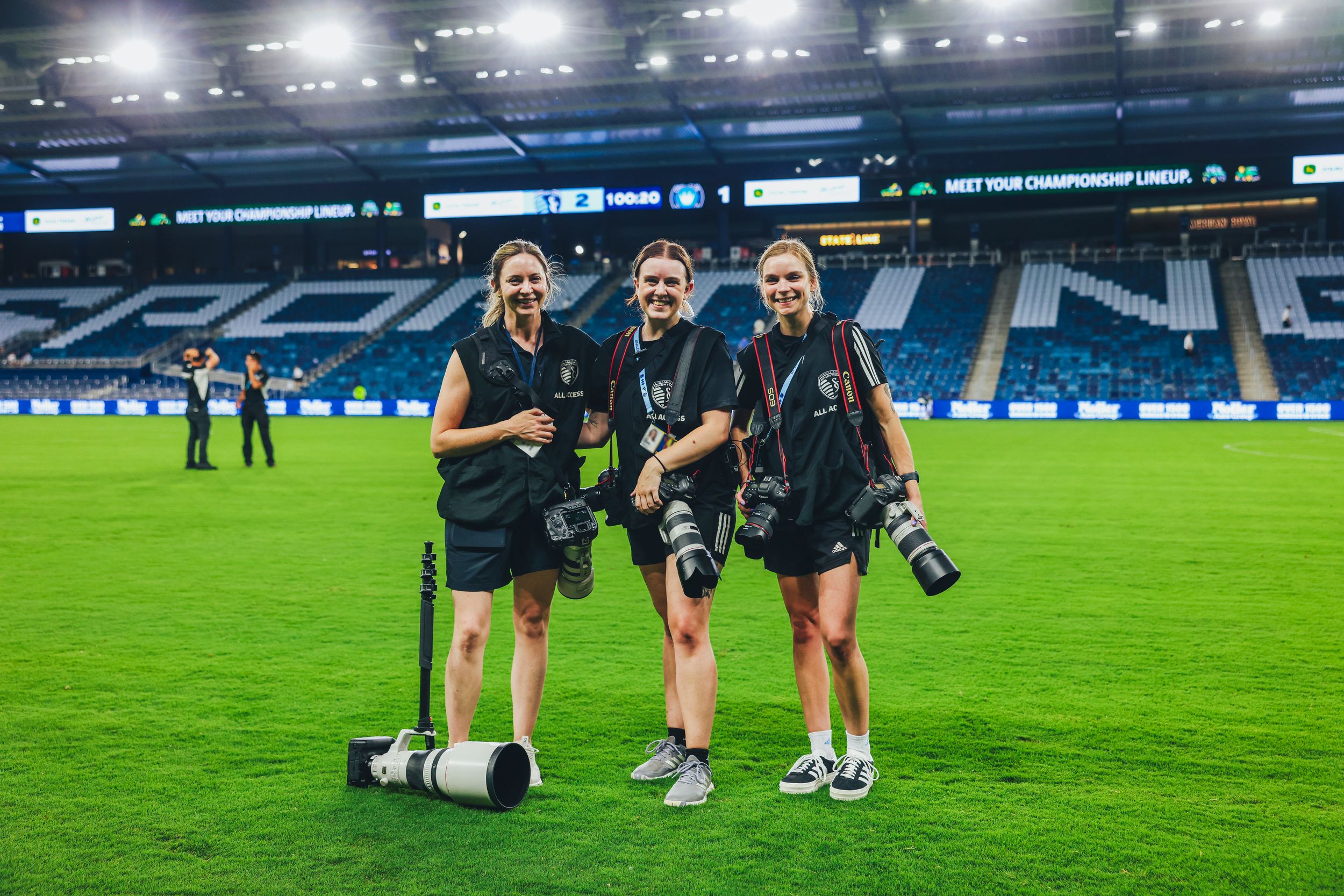 Three female sports photographers smile on a soccer field holding professional cameras and telephoto lenses.