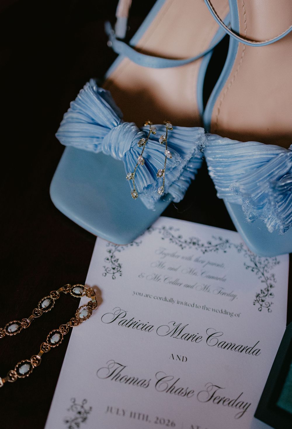 Blue satin wedding heels with bow detail beside a wedding invitation for Patricia Marie Camarda and Thomas Chase Tereday.