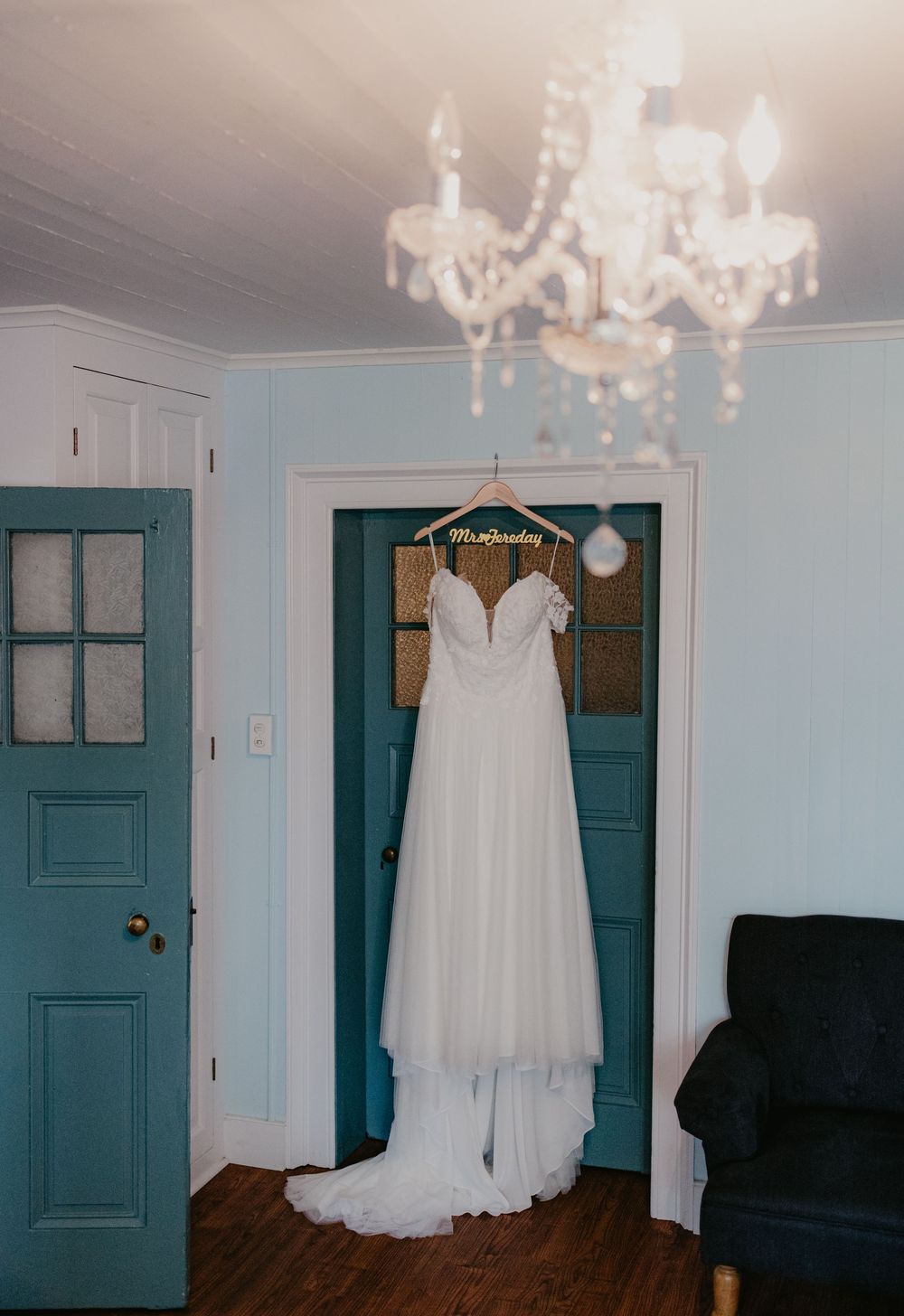 White wedding dress hanging on a personalized hanger beneath a crystal chandelier in a room with teal doors.