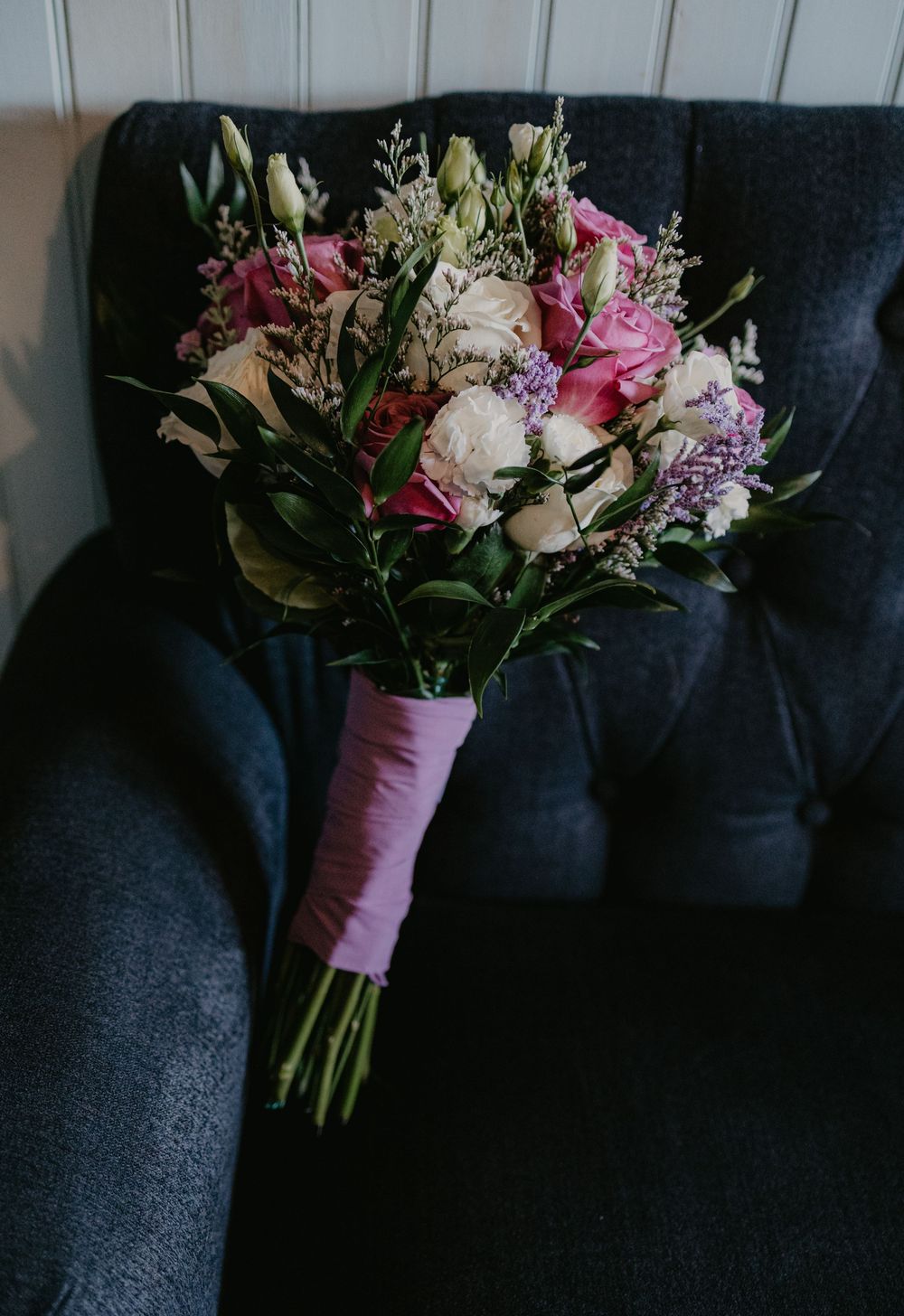 A bridal bouquet with pink roses, white blooms, and greenery wrapped in pink ribbon resting on a dark velvet chair.