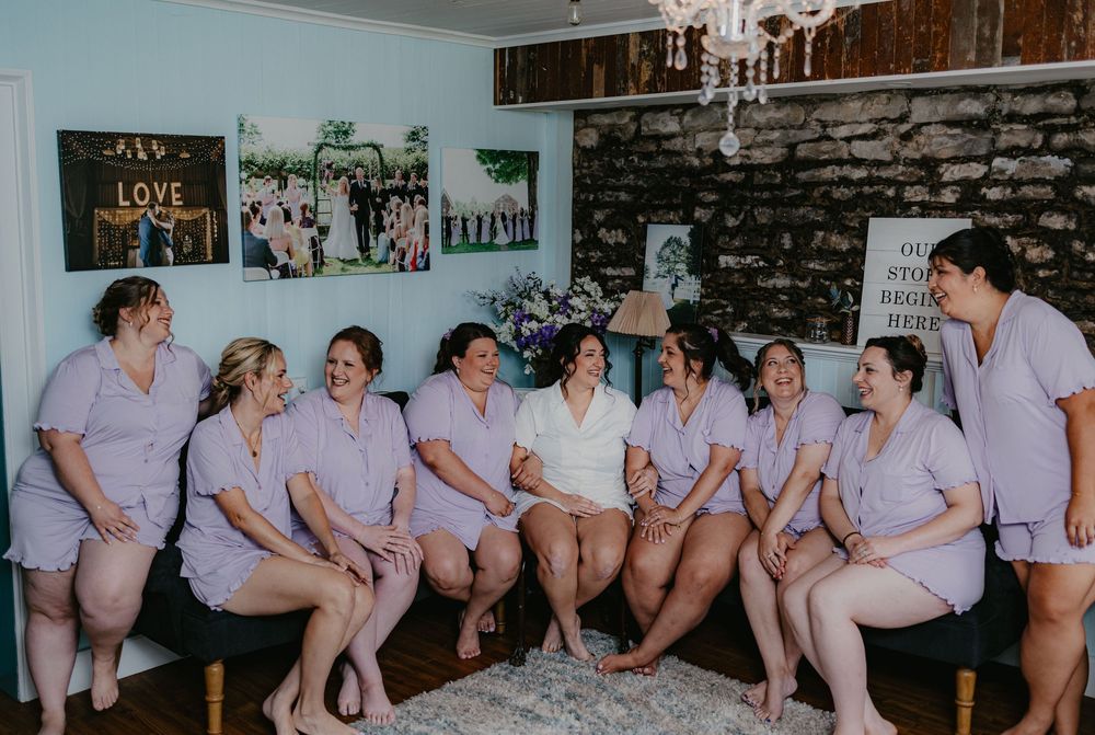 Bride in white surrounded by bridesmaids in lavender matching sets, laughing together in a cozy room before the wedding.