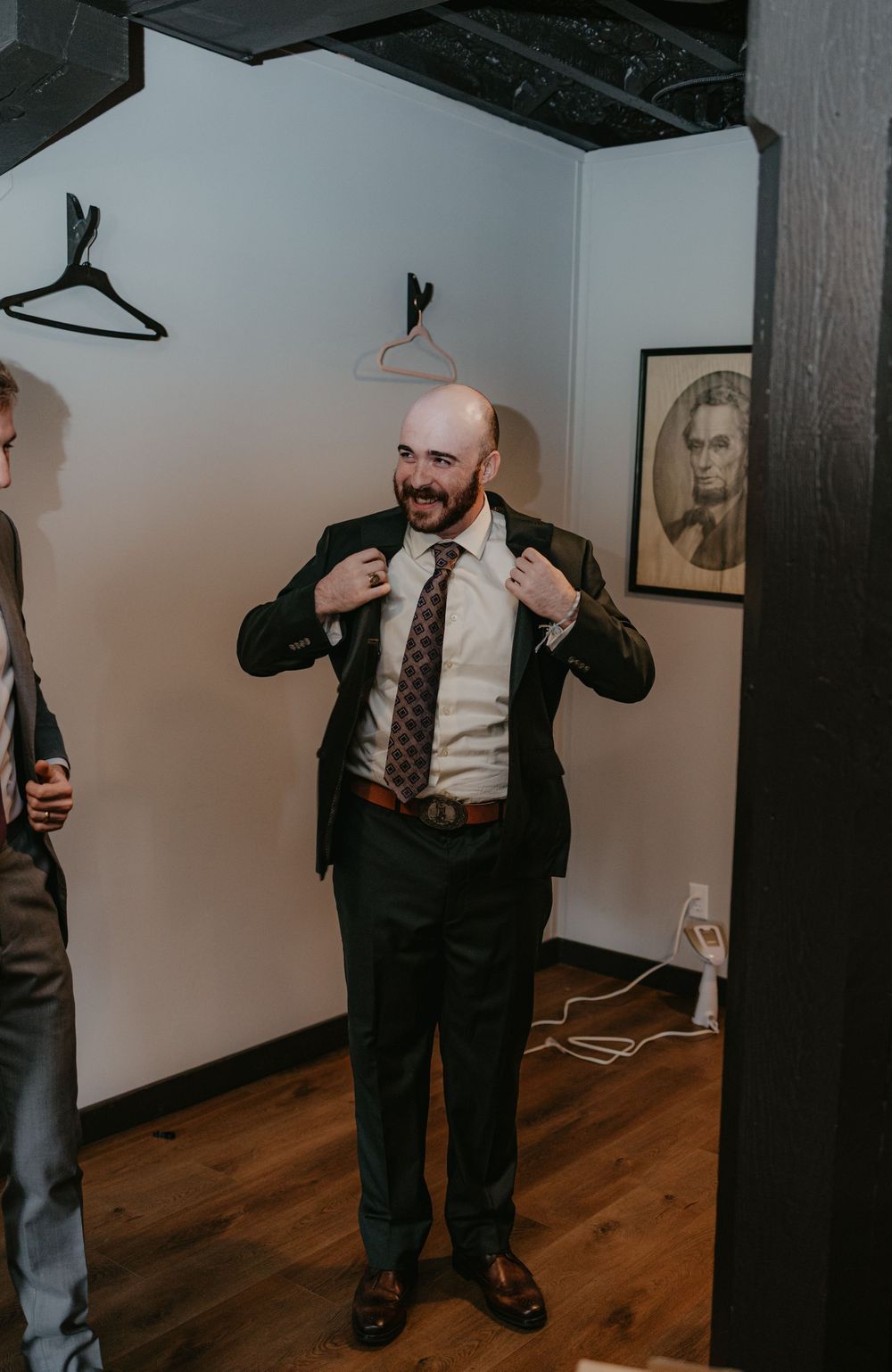Bald man in dark suit and patterned tie adjusting jacket lapels, smiling in a dressing room with portrait art on wall.