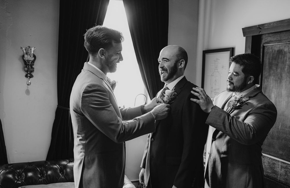 Black and white photo of groomsmen helping groom adjust his boutonniere before wedding ceremony.