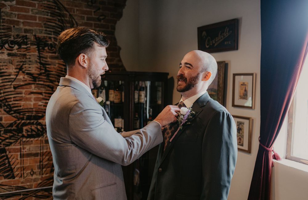 A groomsman adjusts the bow tie of a bearded groom in a suit inside a rustic brick-walled venue.