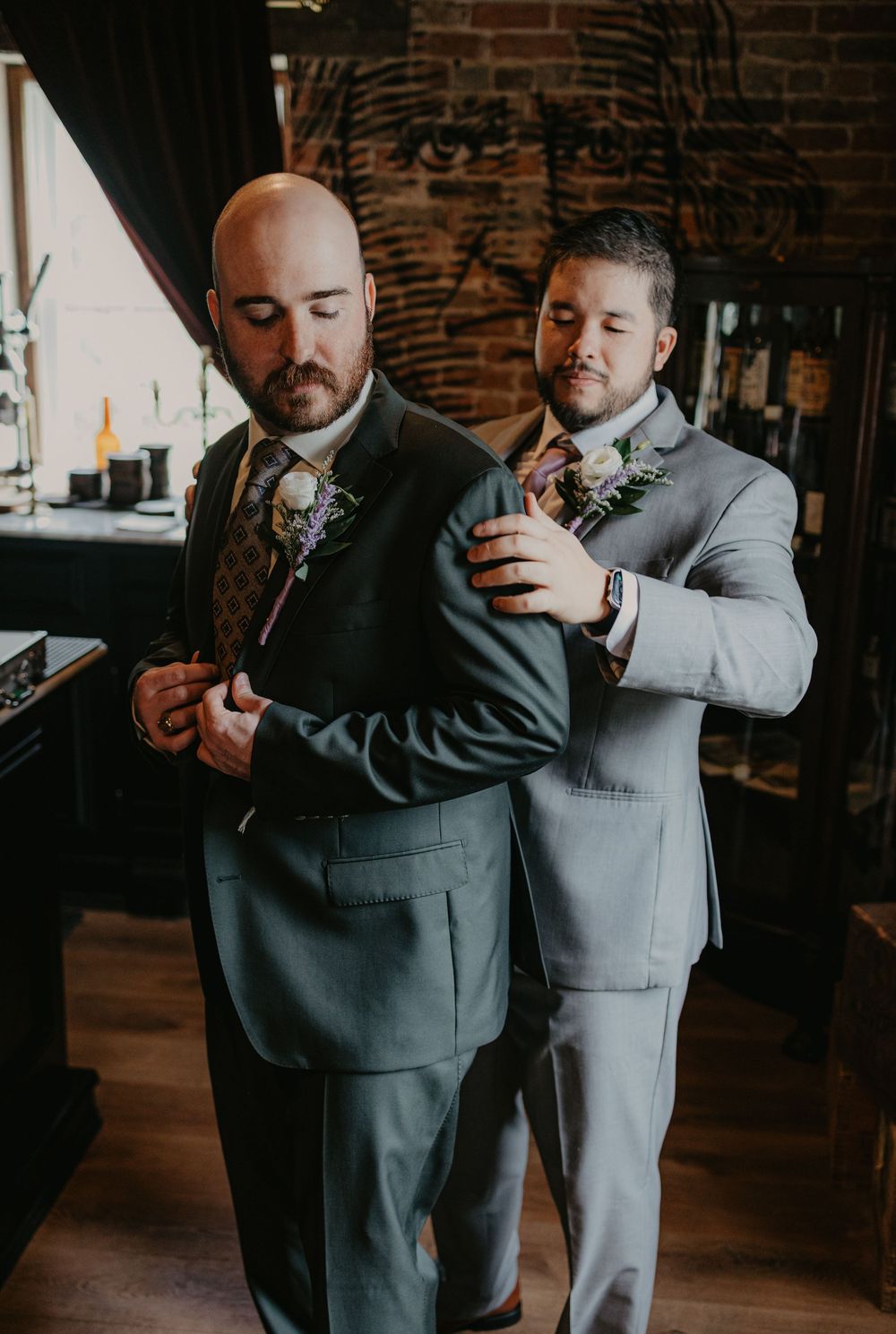 Two grooms in gray suits help each other get ready on their wedding day in a rustic venue.