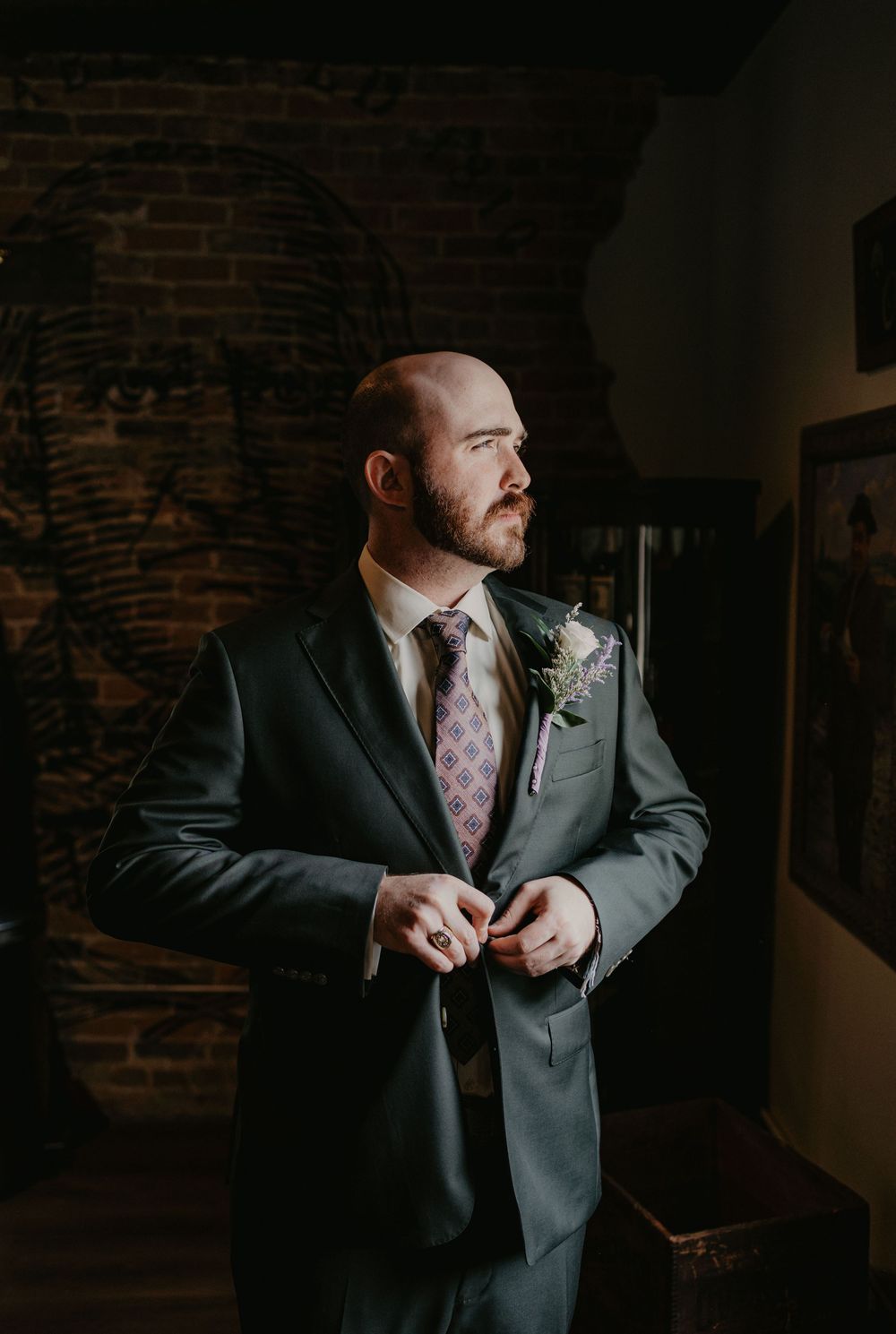 Groom in gray suit with floral tie and boutonniere buttoning jacket in moody brick-walled venue.