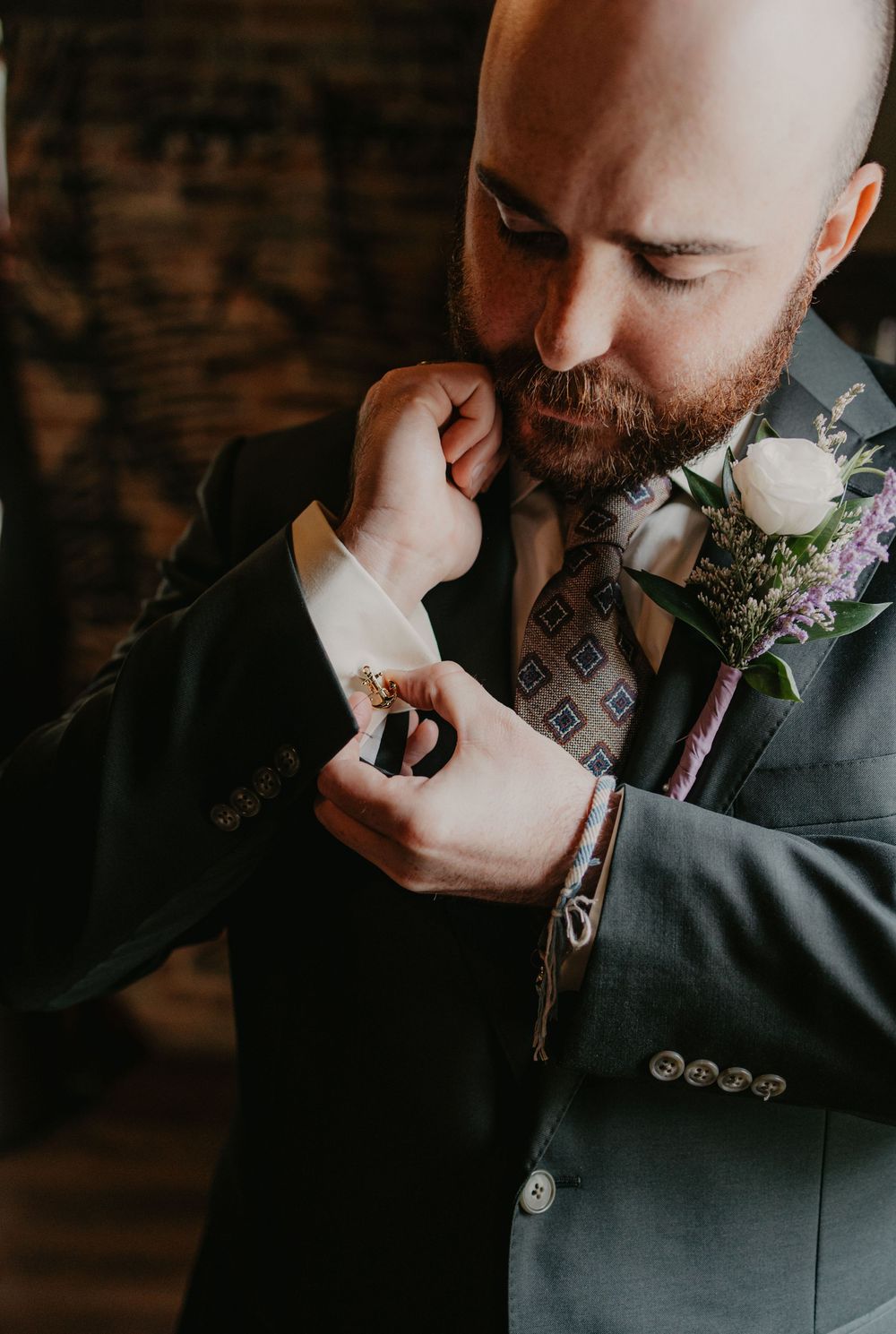 A dapper groom in a gray suit fastens his cufflinks, wearing a floral boutonniere and patterned tie on his wedding day.